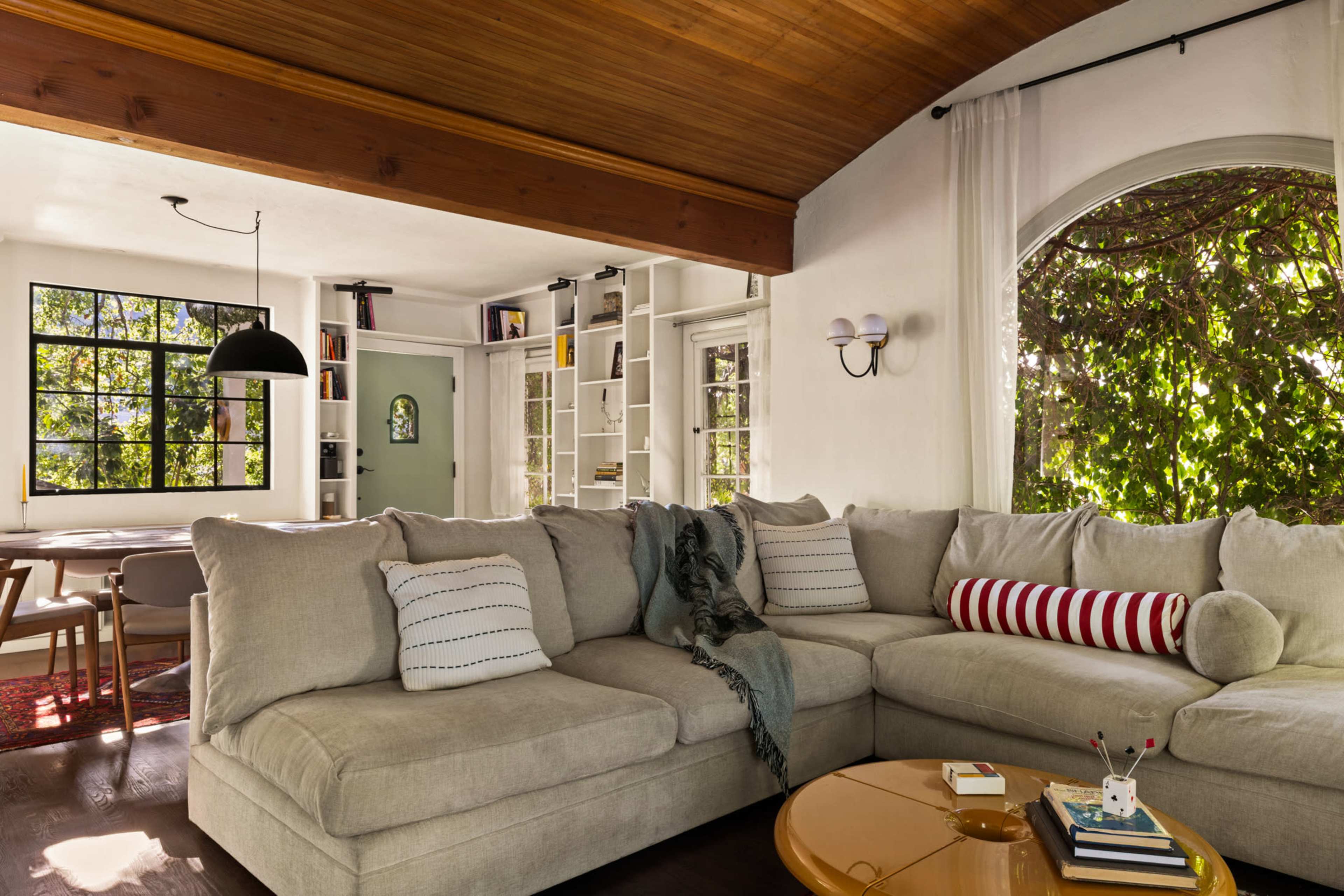 The image shows a cozy living room with a large, L-shaped beige sectional sofa, a round coffee table, and large windows that allow natural light to fill the space.