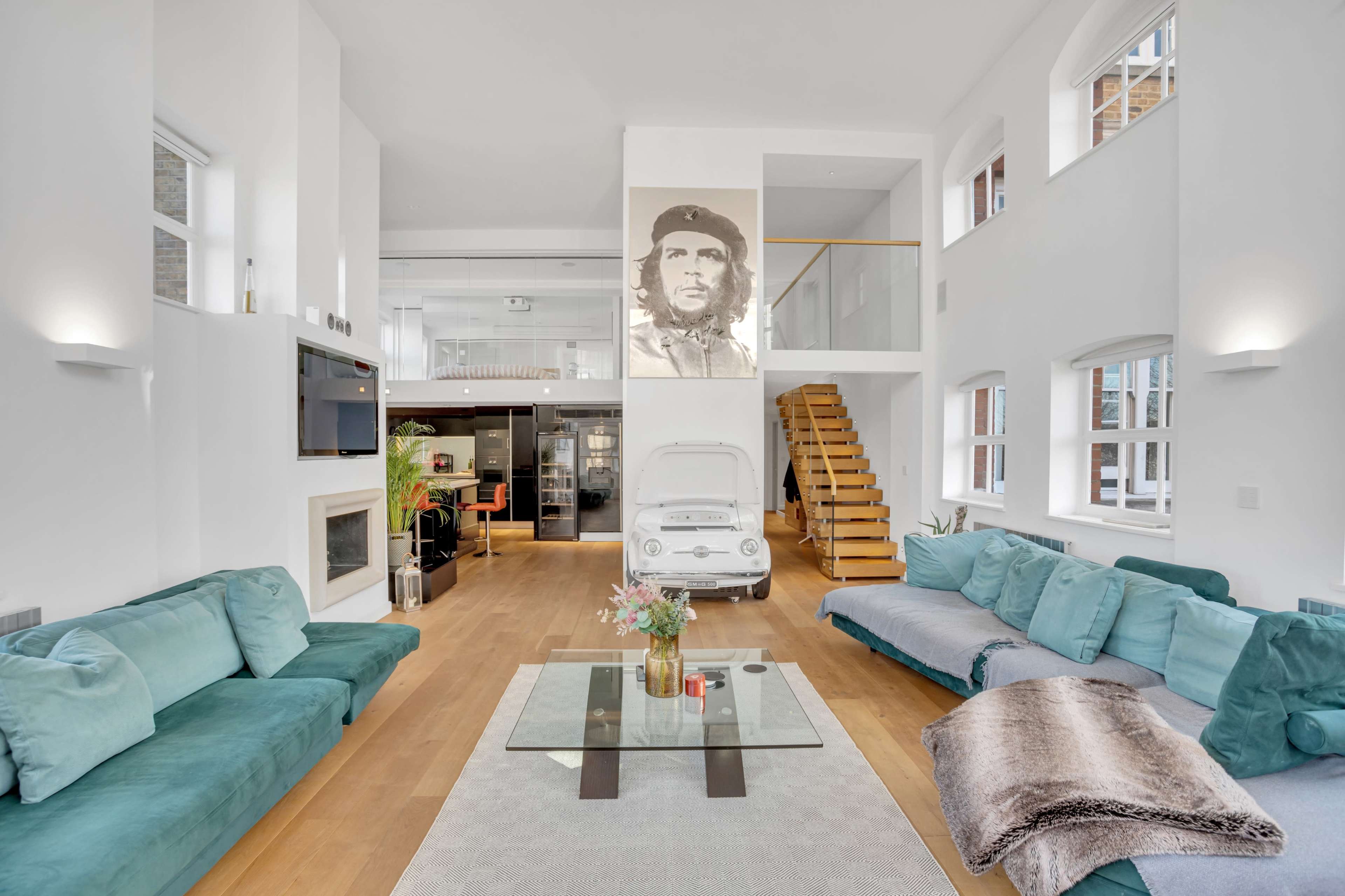 The image shows a modern living room with two teal sofas, a glass coffee table, and a staircase leading to an upper level, featuring a large portrait of Che Guevara on the wall.