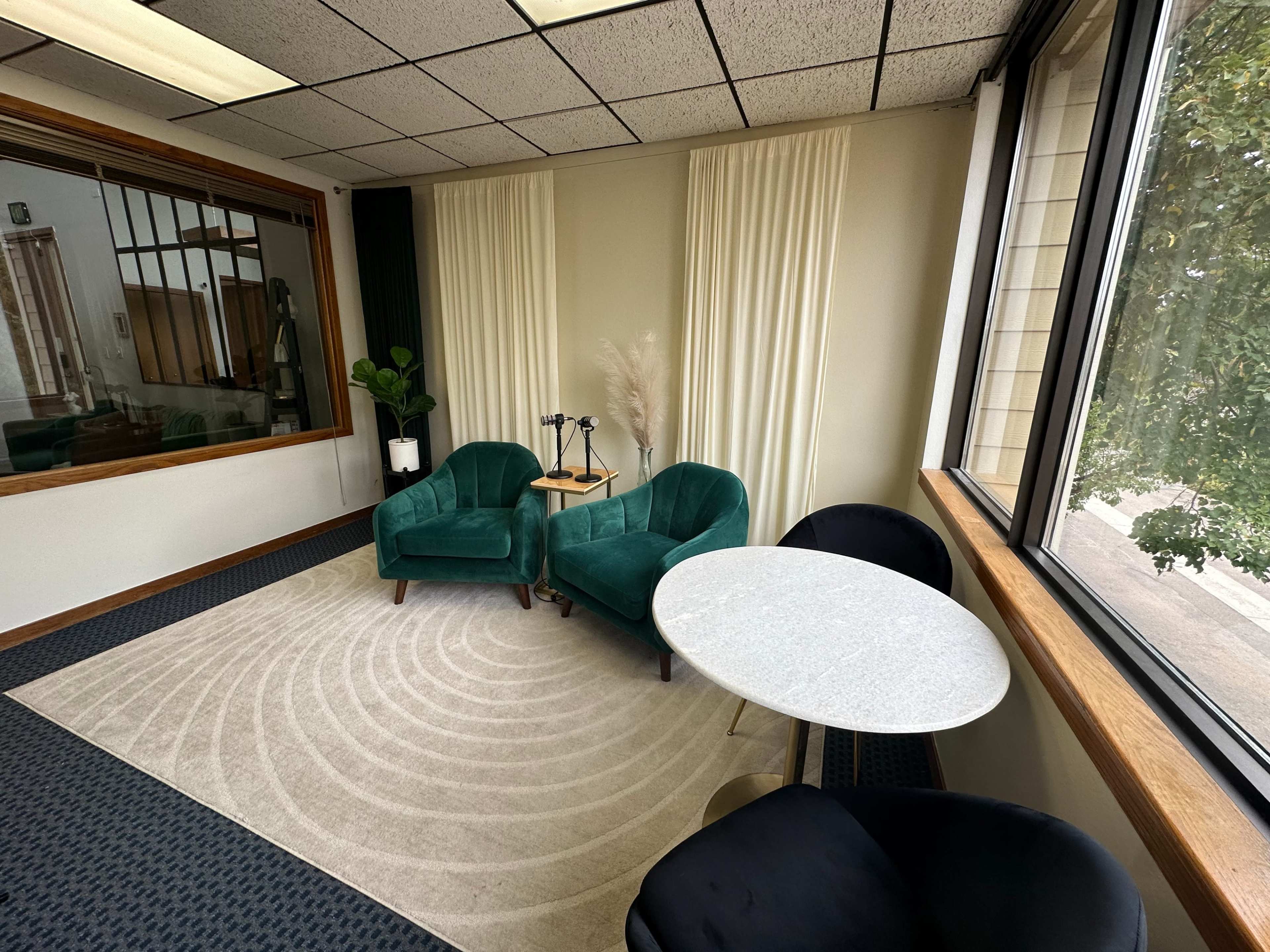 The image shows a small, well-lit waiting area featuring two green velvet chairs, a round marble-top table, and a large window with sheer curtains.