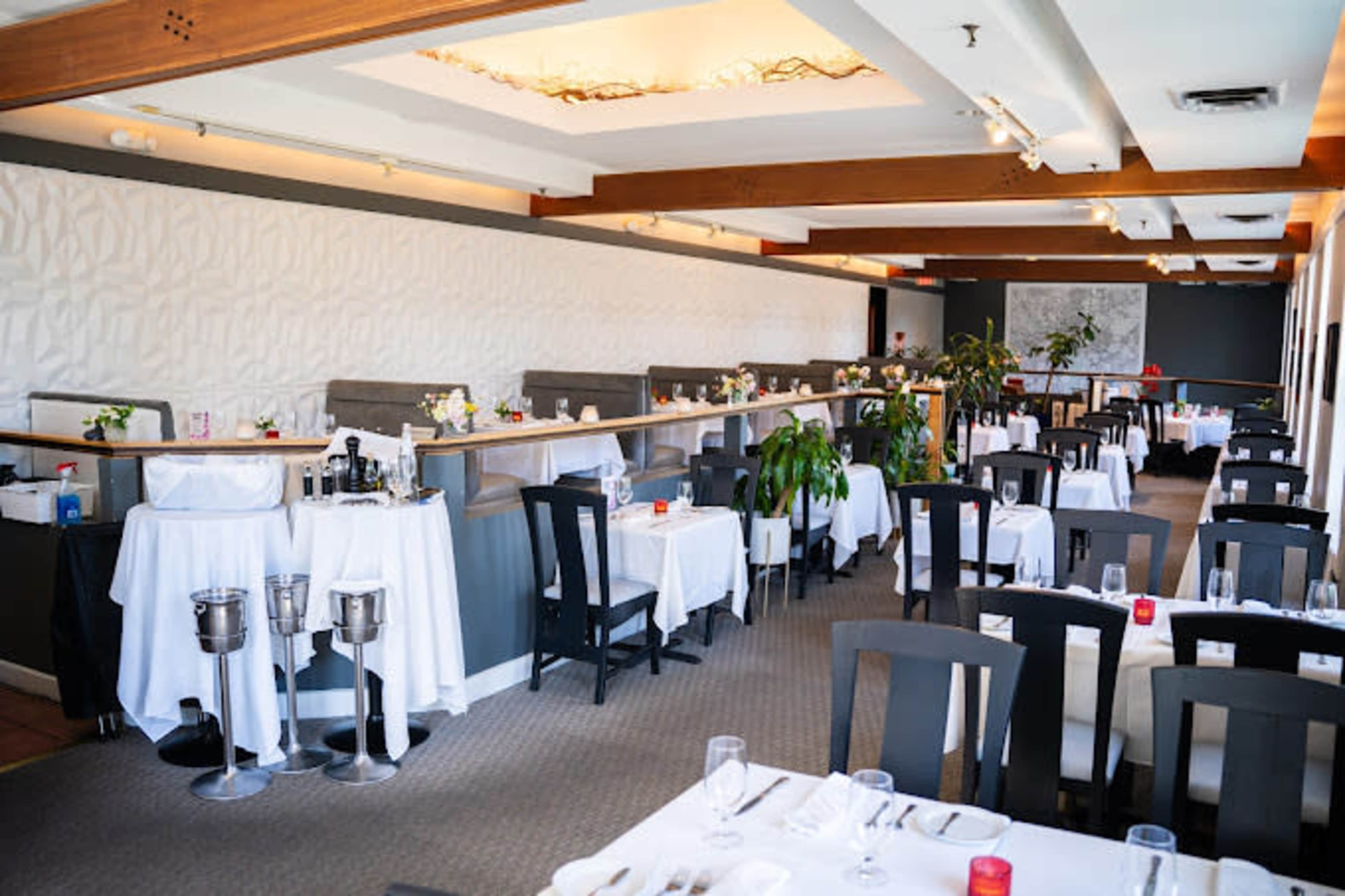 The image shows an elegantly arranged restaurant interior with neatly set tables, white tablecloths, and a contemporary design featuring wooden beams and plants.