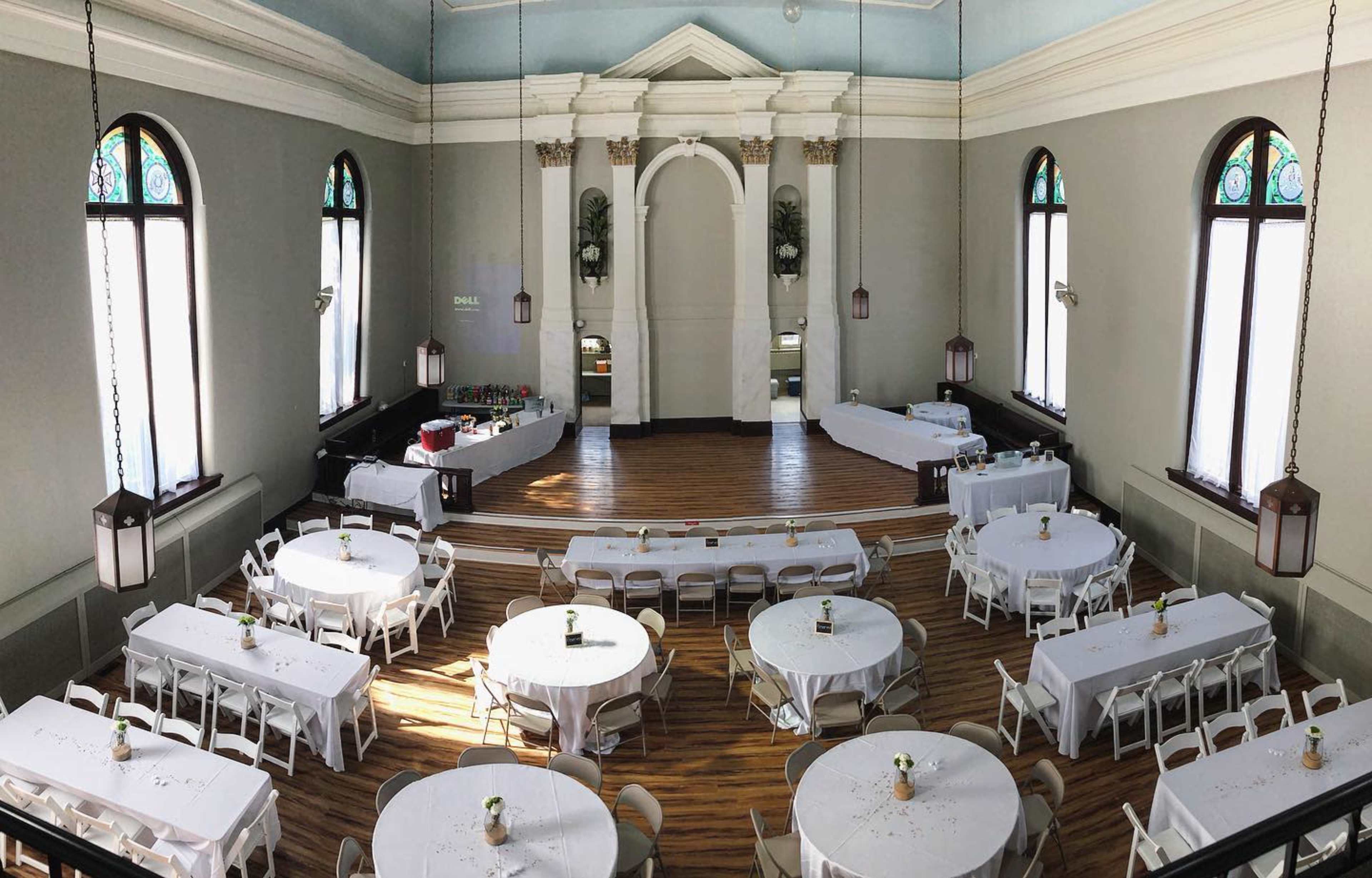 The image shows a spacious, well-lit hall with round tables set up and a raised area in the background that appears to be a stage or serving area.