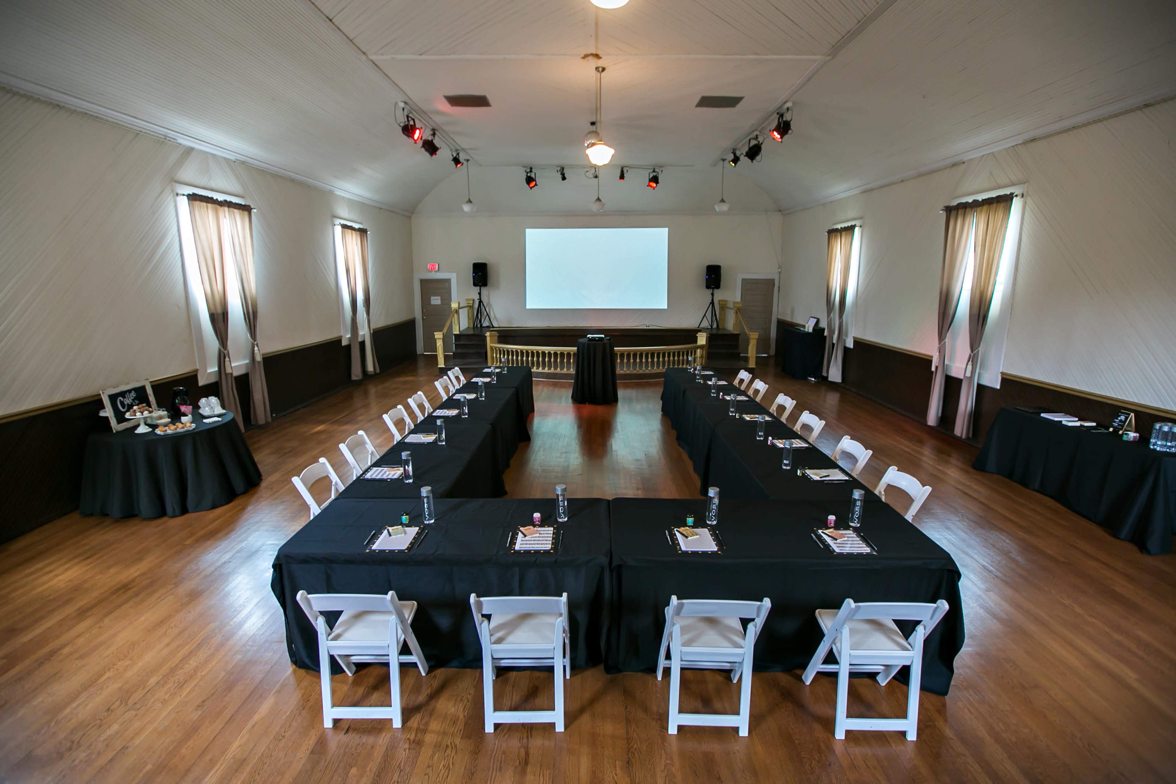 A spacious conference room is set up with tables arranged in a U-shape, surrounded by chairs, and equipped with a projector screen at one end.