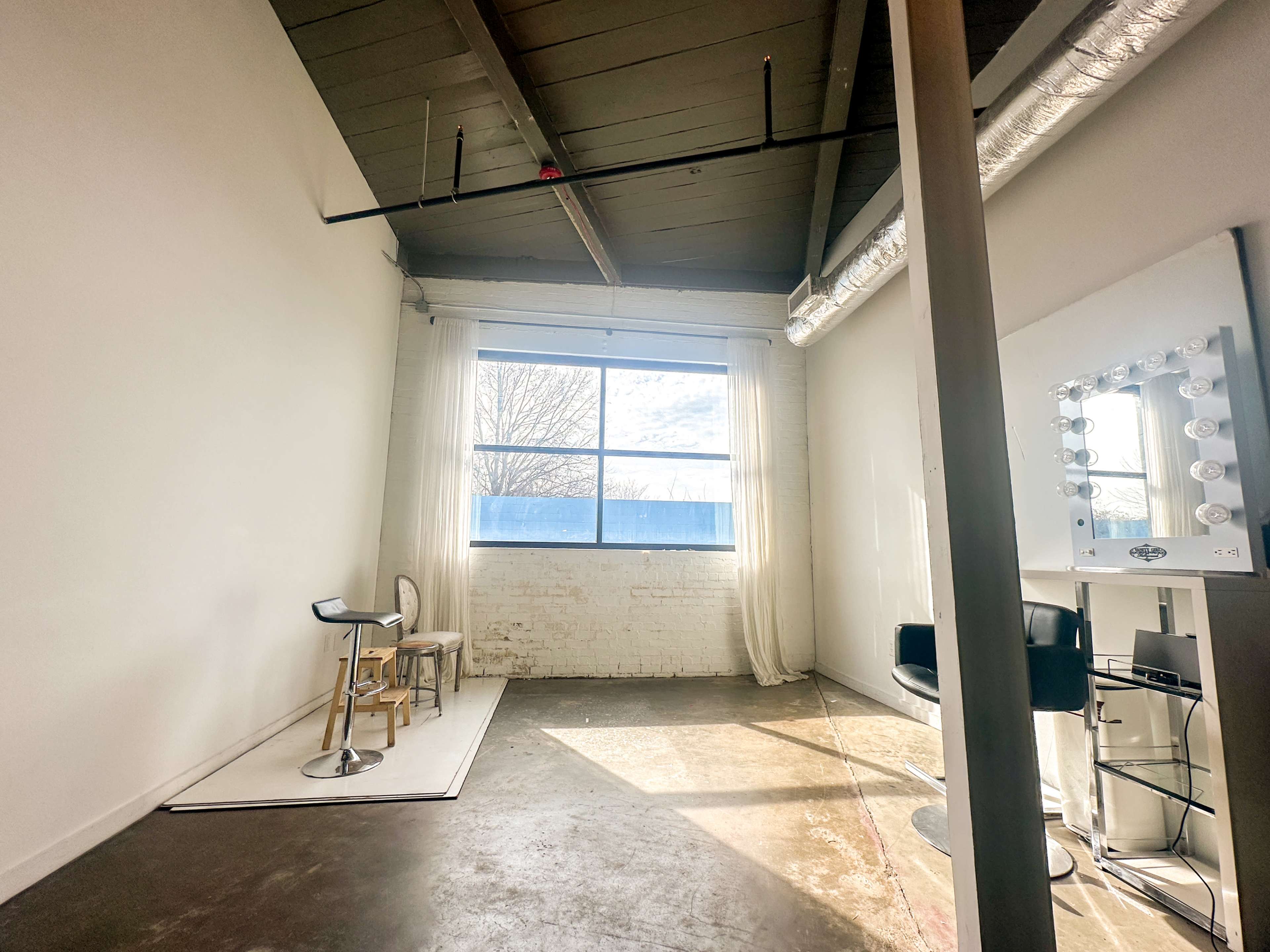 The image shows a spacious, empty room with large windows, a few pieces of furniture, and a makeup mirror against the wall.