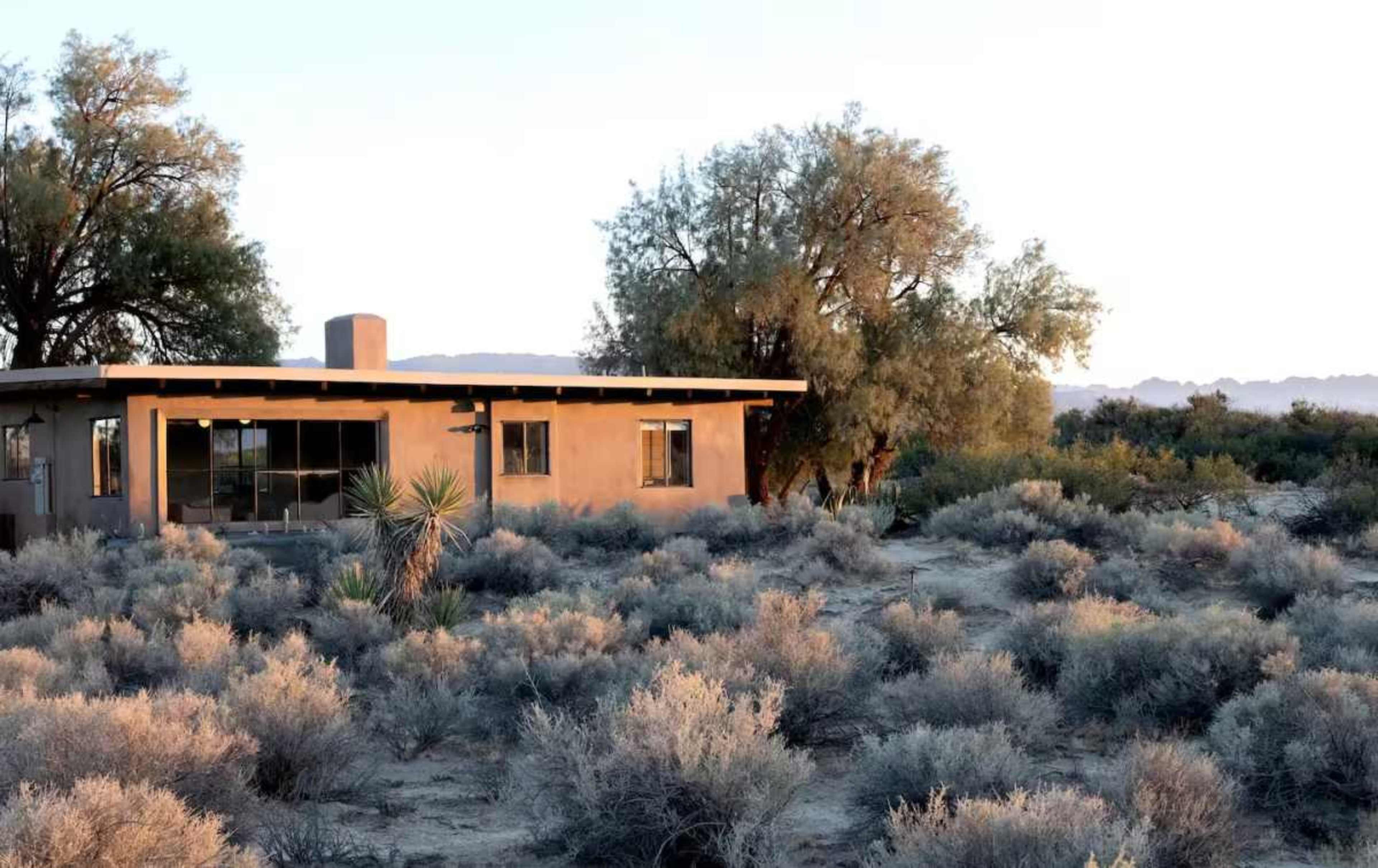 A modern adobe-style house is set amidst low desert vegetation and sparse trees in a dry landscape.