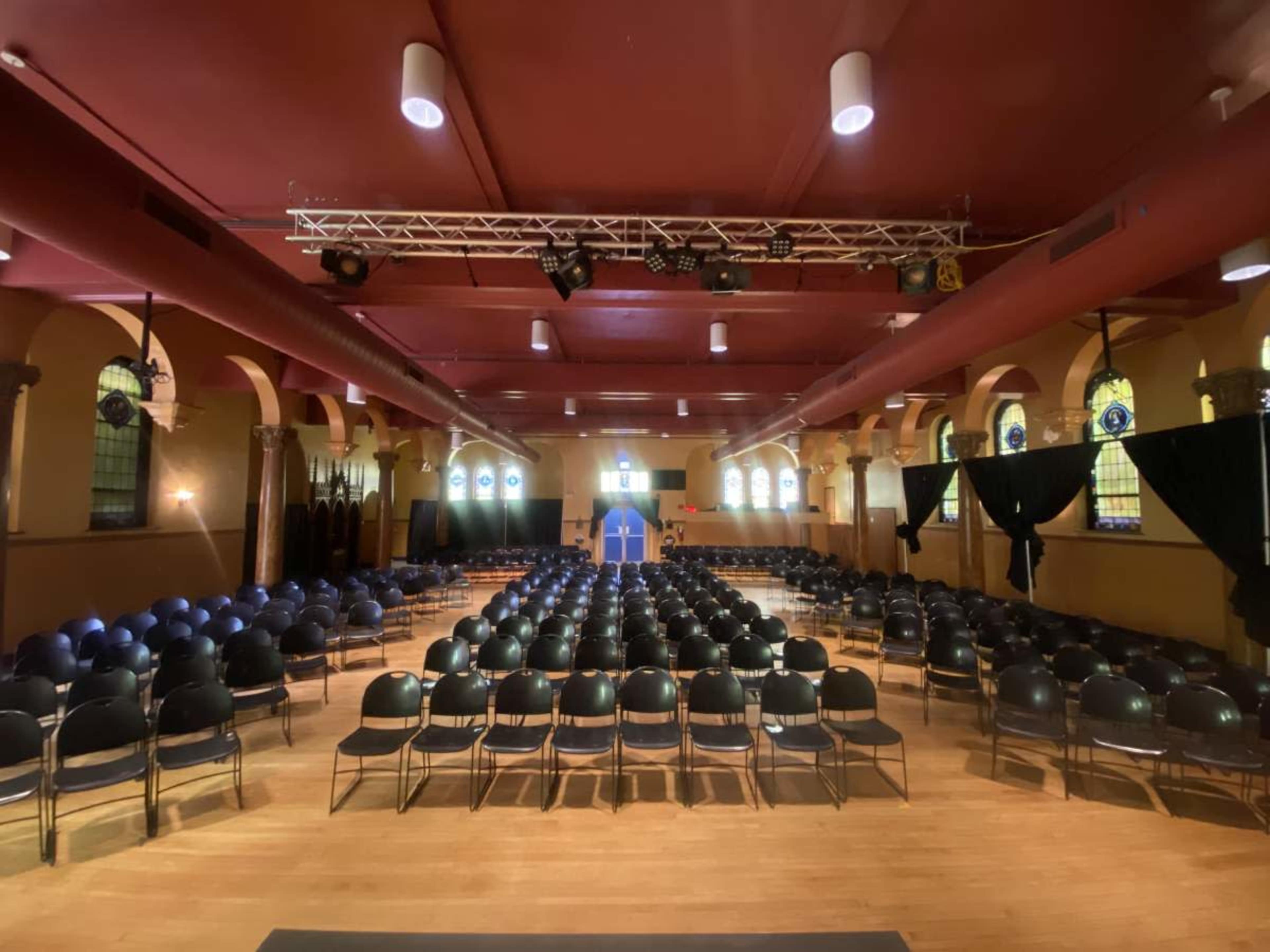 A large hall set up with rows of black chairs facing a stage at the front, with stained glass windows lining the walls.