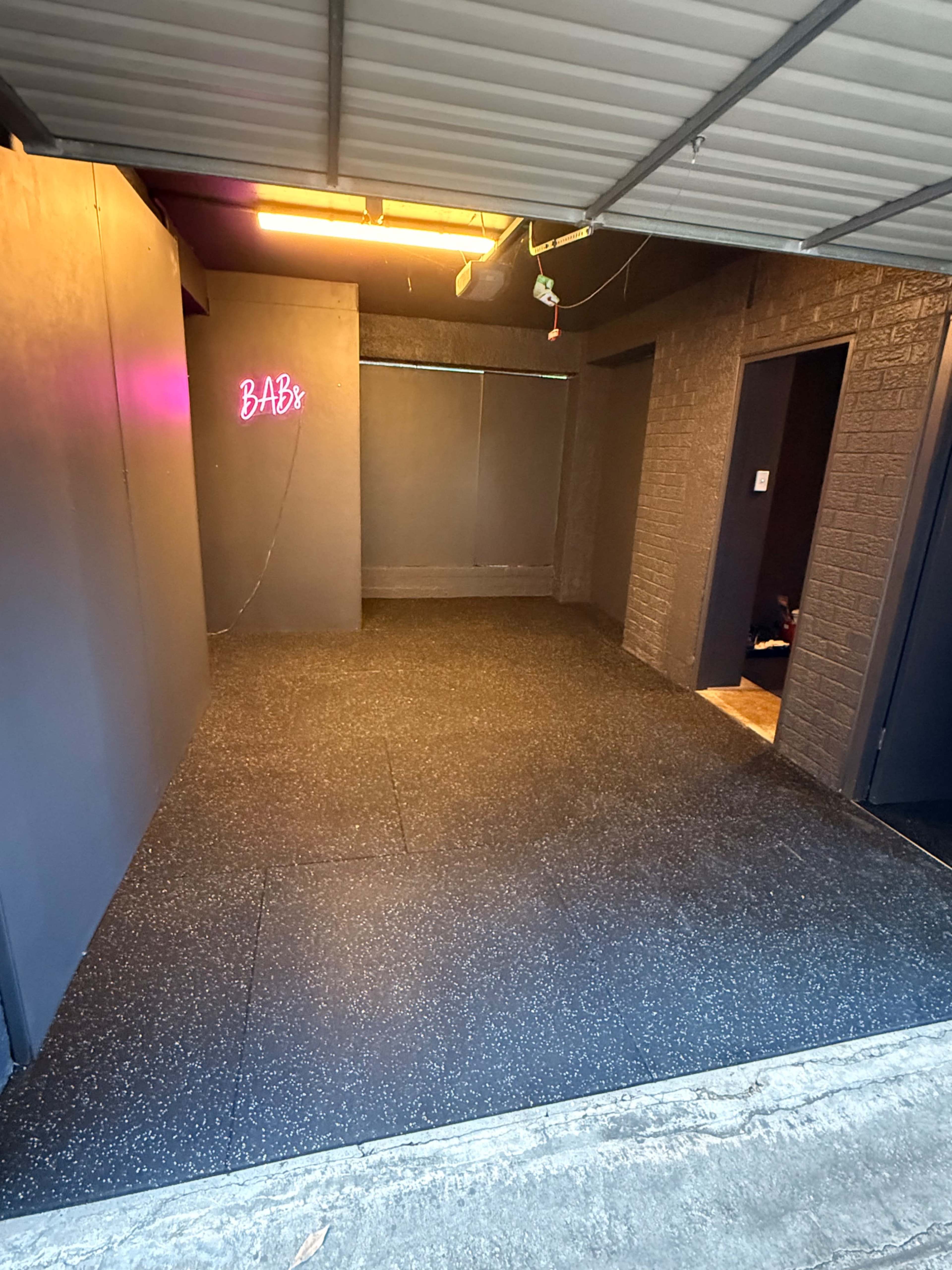 The image shows a dimly lit indoor space with gray walls, a black rubber floor, and a neon sign that reads "BABY."