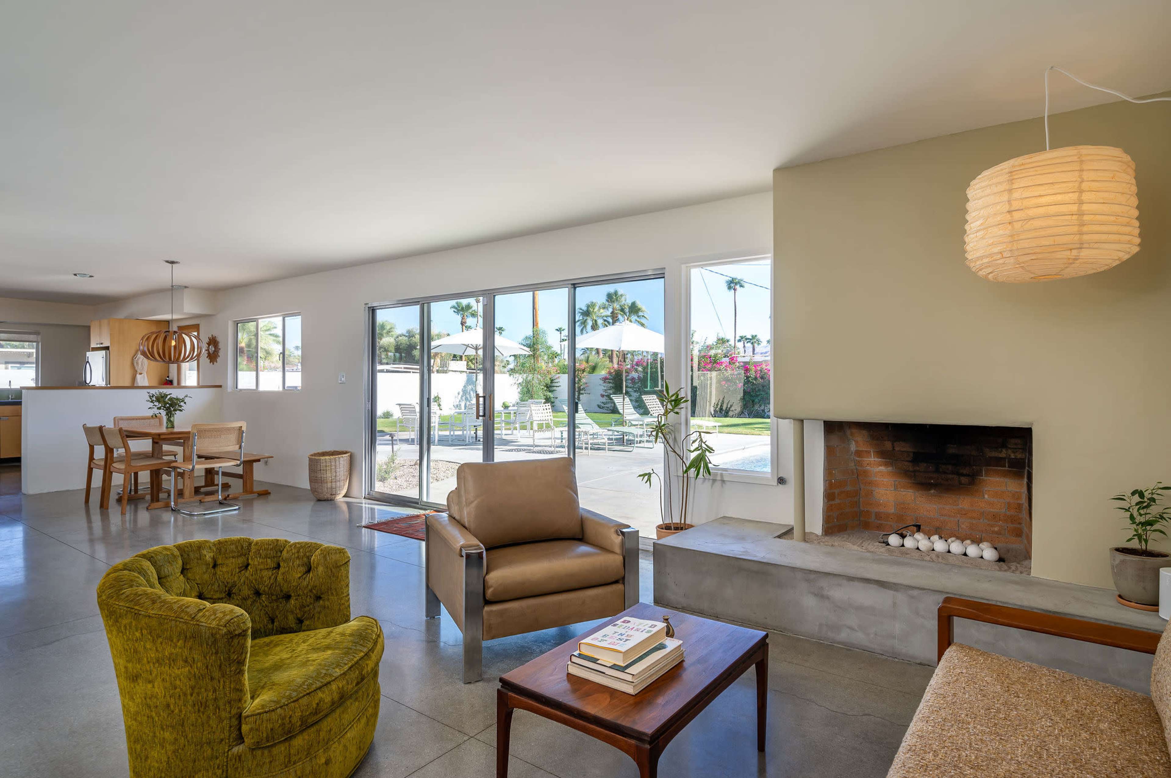 The image shows a modern living room with a yellow armchair, brown sofa, and a fireplace, featuring large glass doors that open to a patio with a pool and palm trees in the background.