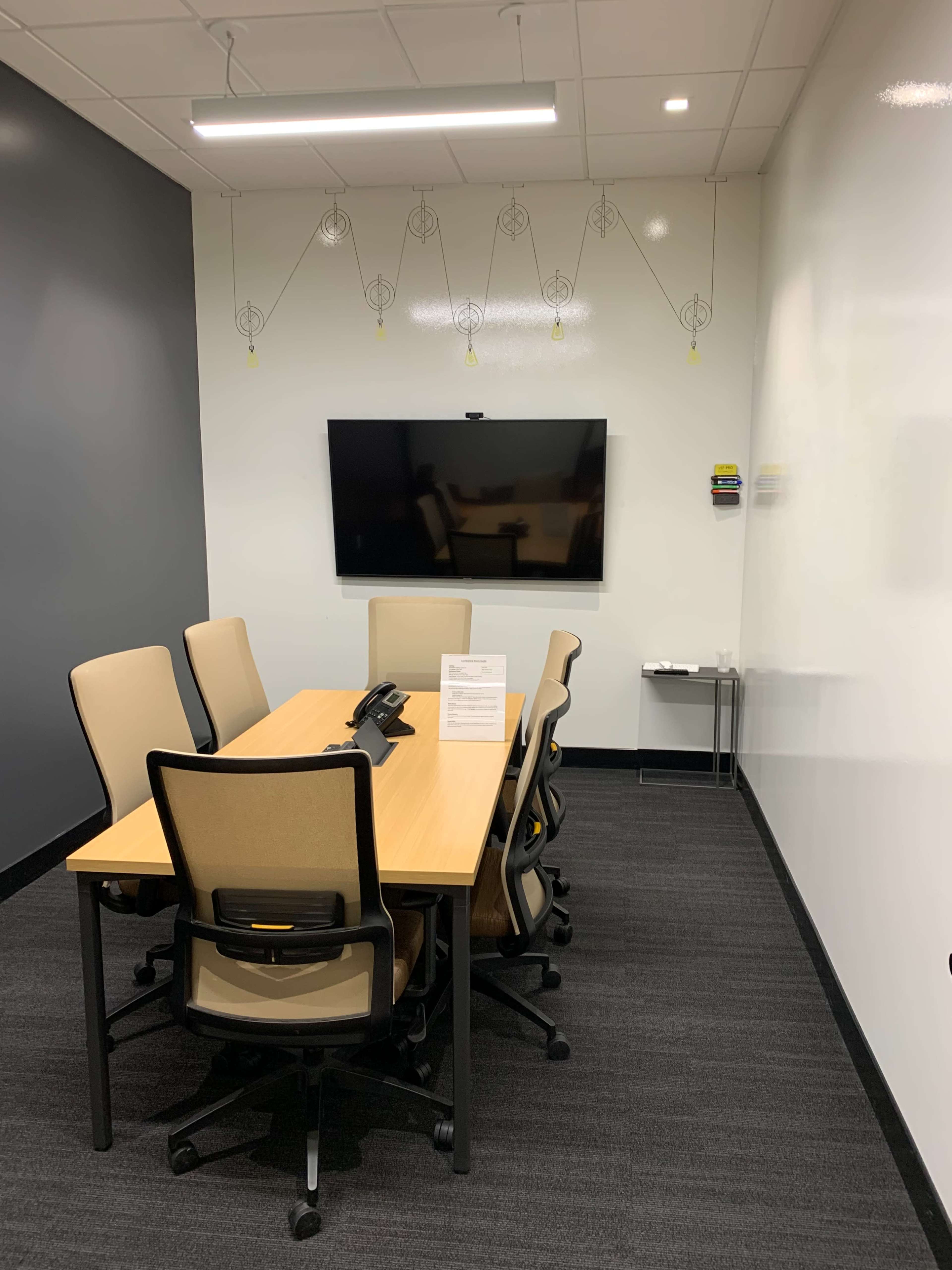 The image shows a small conference room with a rectangular table surrounded by six chairs, a wall-mounted television, and a small side table.