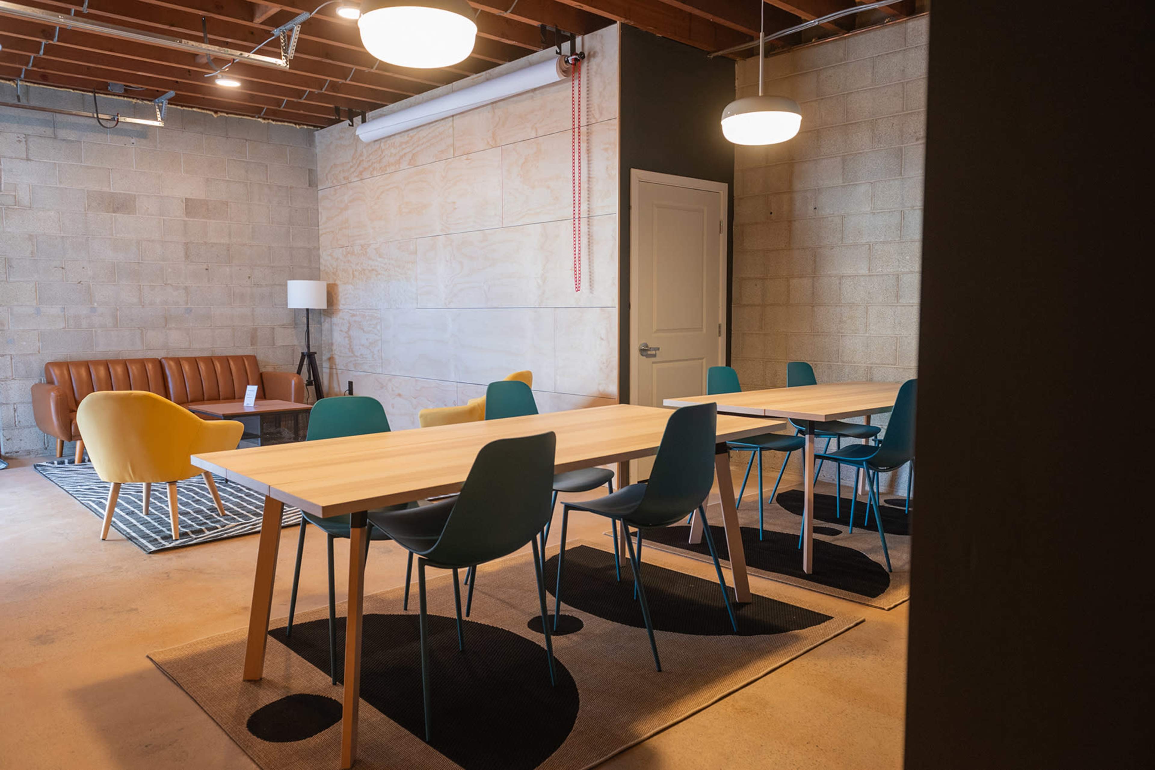 A modern meeting room features a wooden table surrounded by blue and green chairs, with a brown couch and a lamp in the background.