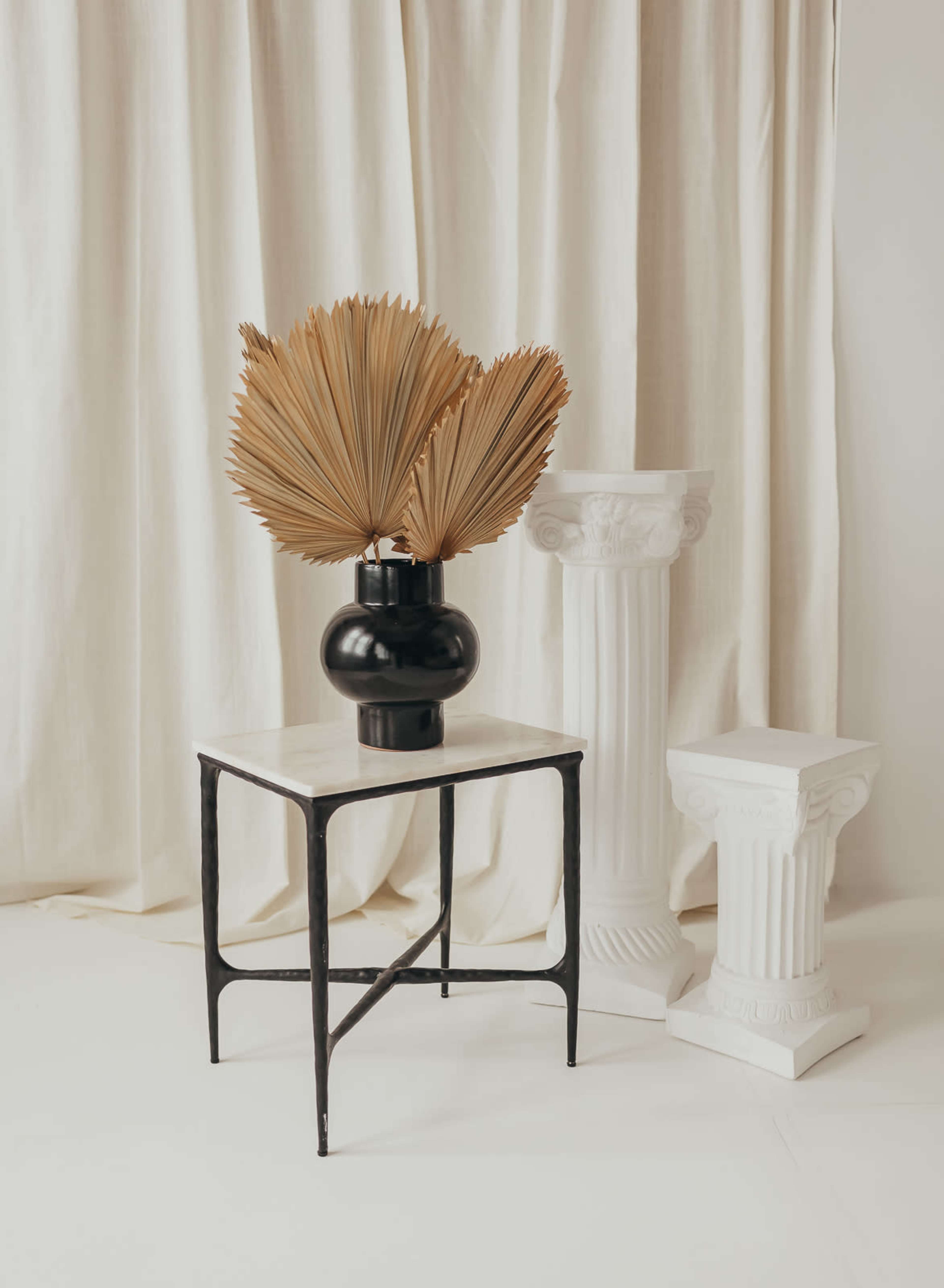 A black vase with dried palm fronds stands on a marble-topped table beside two white columns against a backdrop of cream-colored curtains.