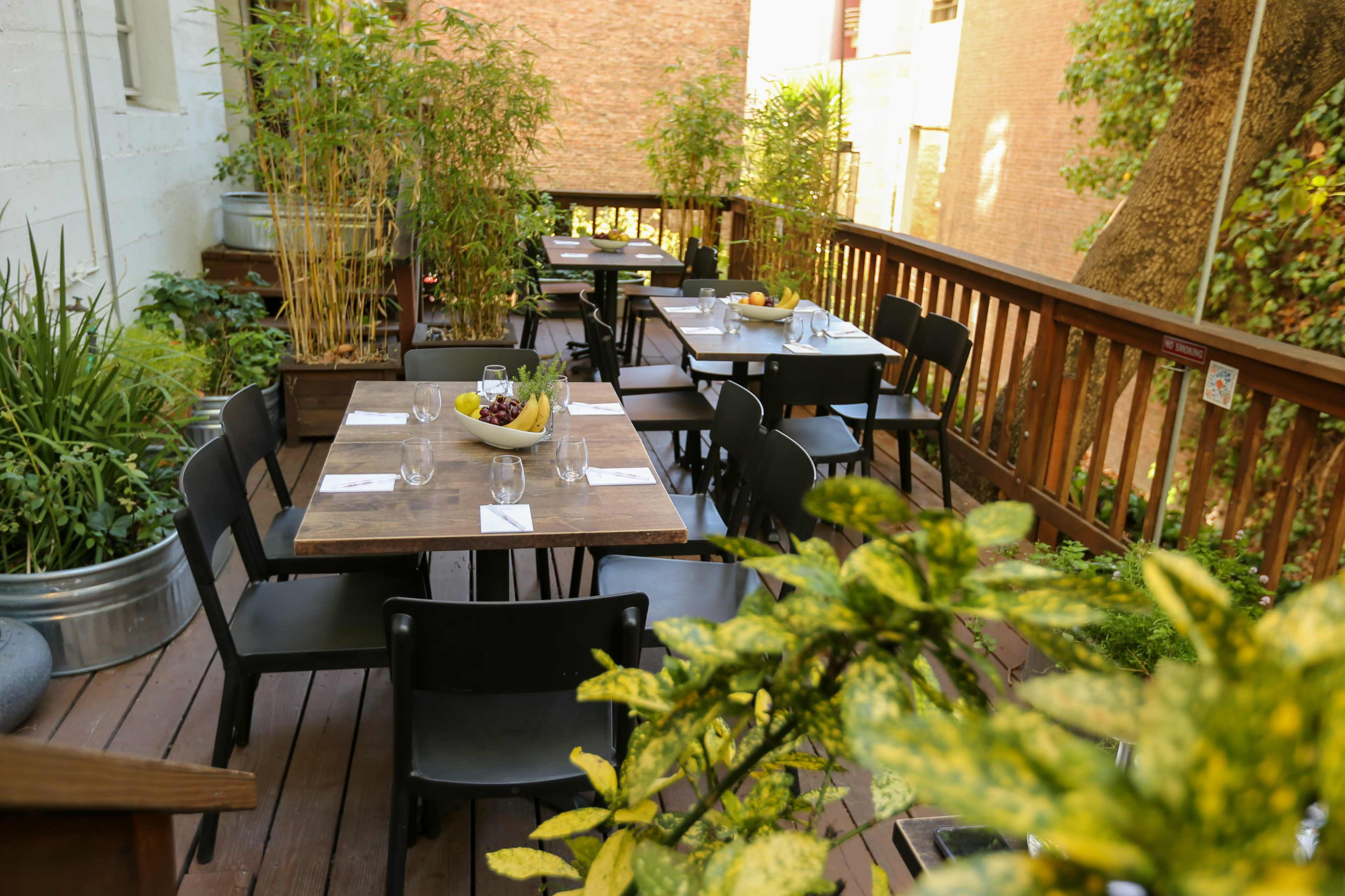 An outdoor dining area with wooden tables set for meals, surrounded by greenery and bamboo plants.