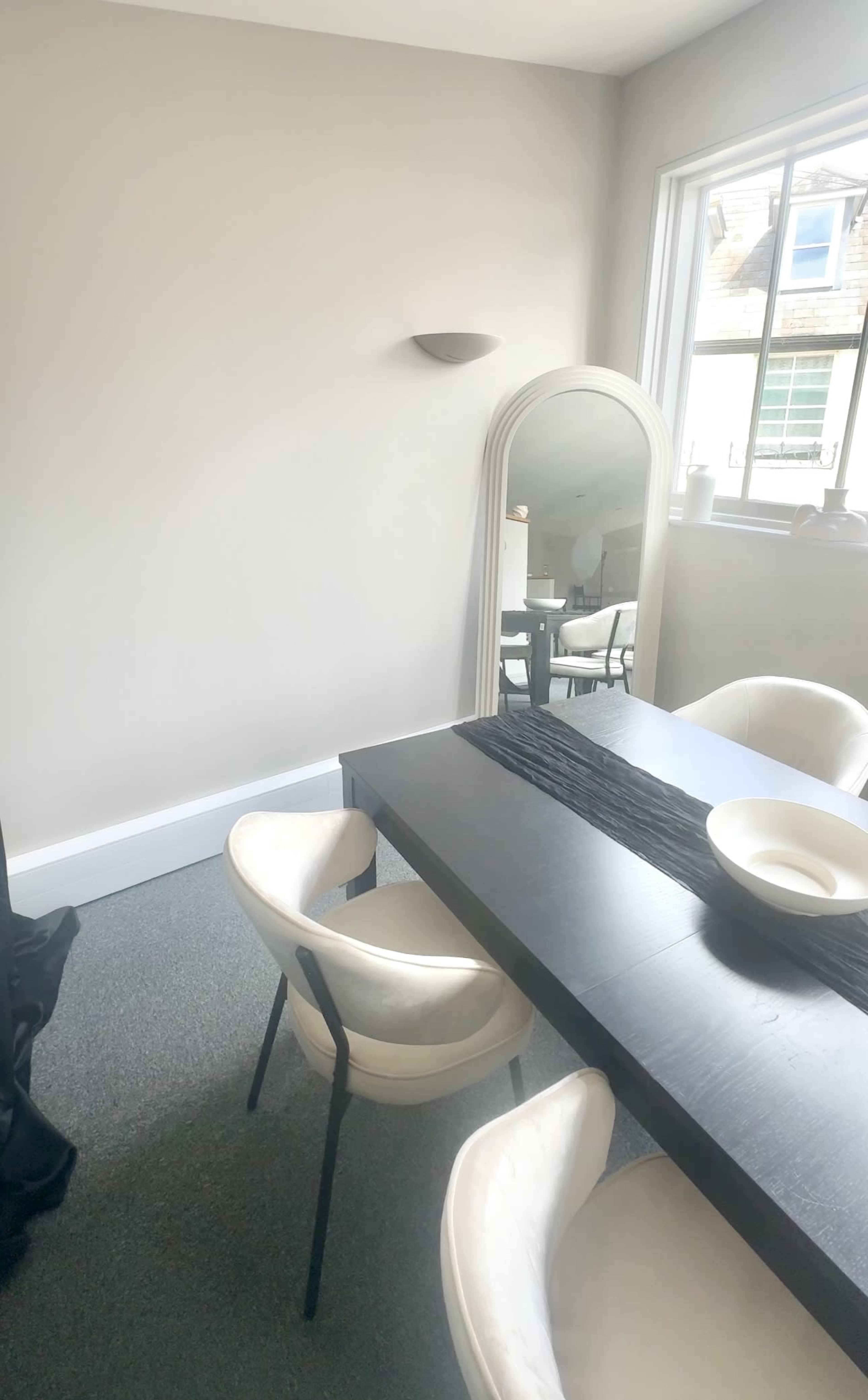 A dining area features a black table, two white chairs, and a large mirror reflecting the light from a window.