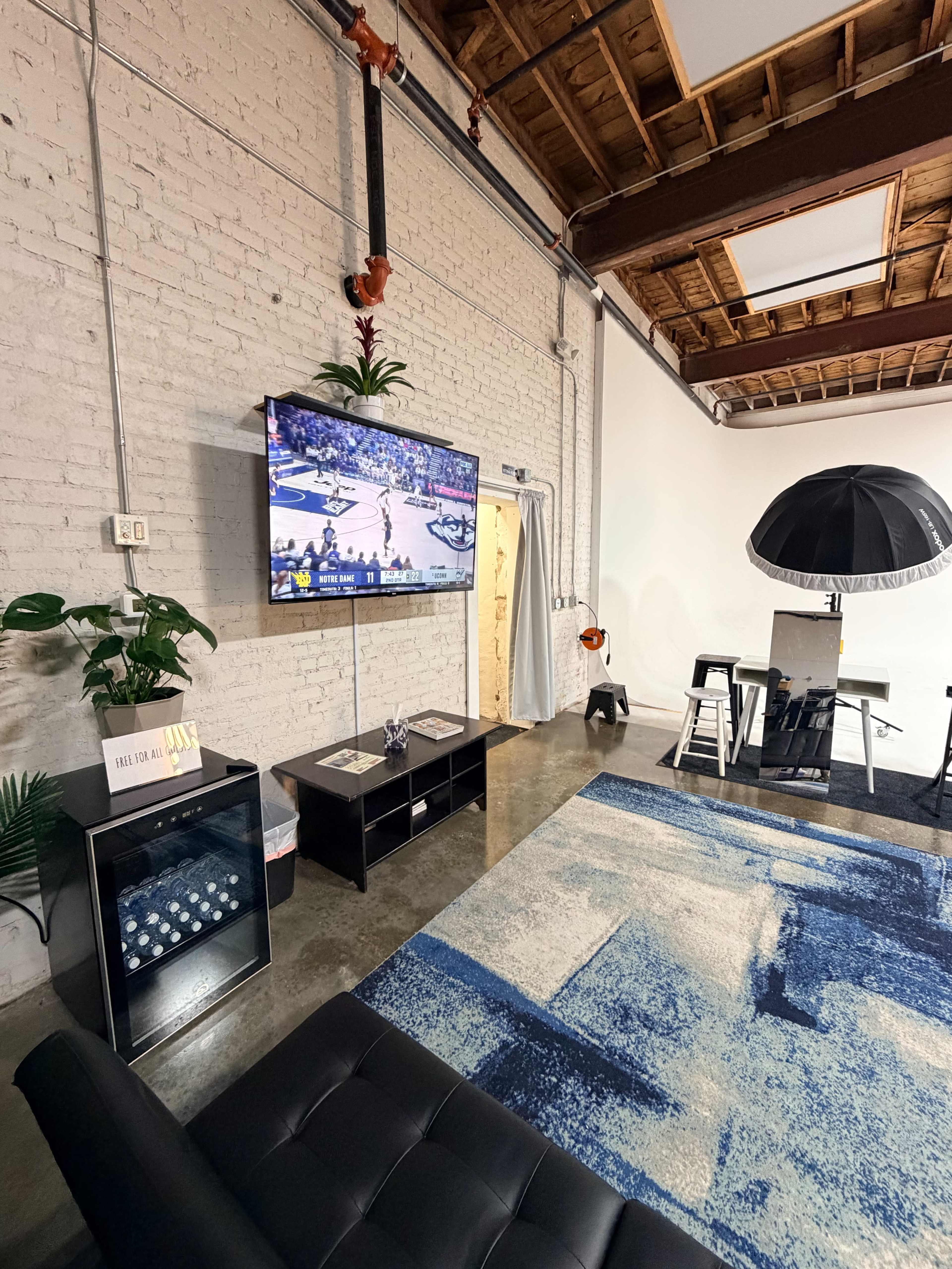 The image shows a modern living space featuring a wall-mounted television, a black sofa, a coffee table, and a rug with blue patterns, all situated against a white brick wall.