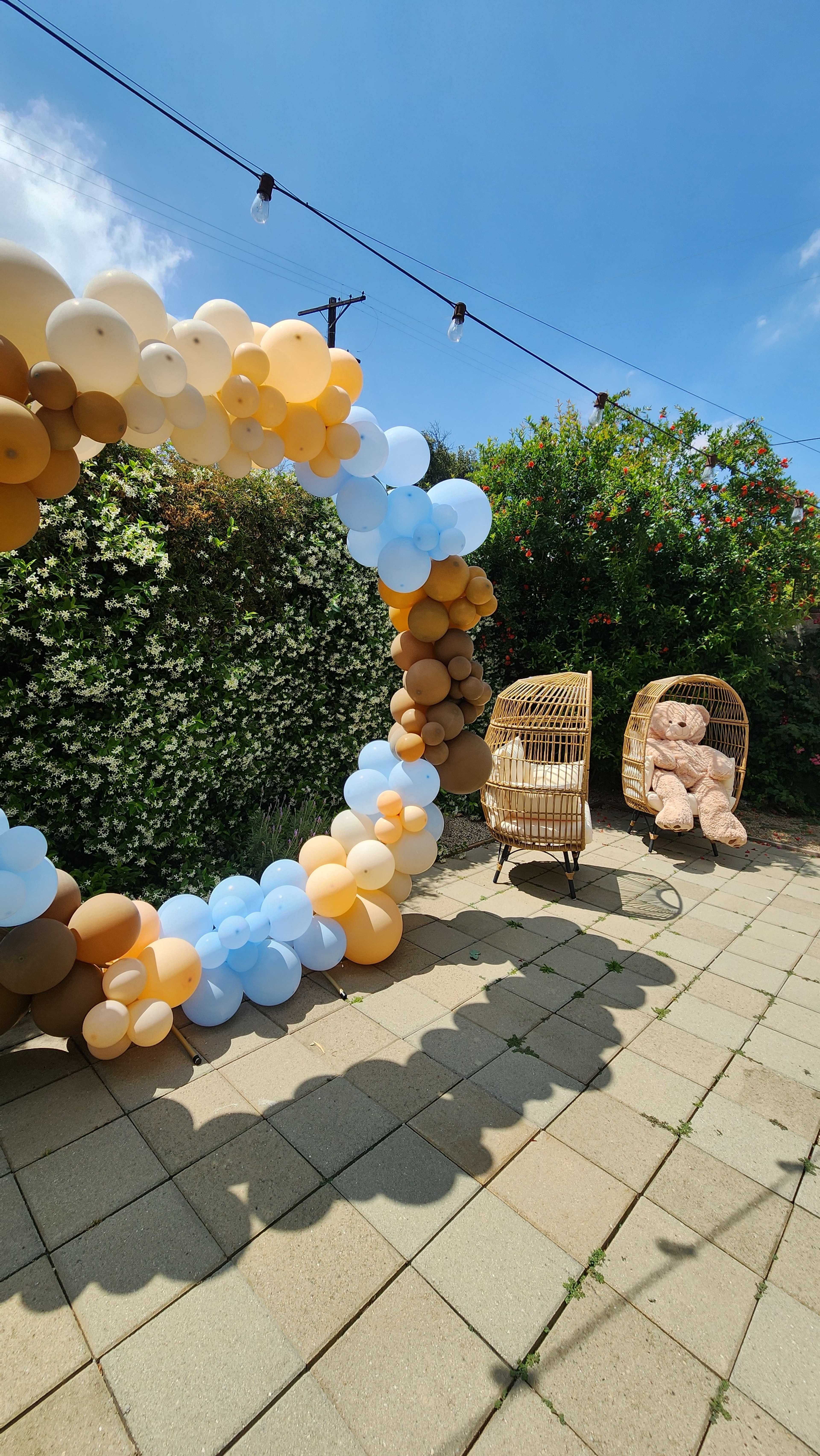A circular balloon arch made of blue, brown, and beige balloons stands next to two wicker chairs and a large teddy bear in a garden setting.