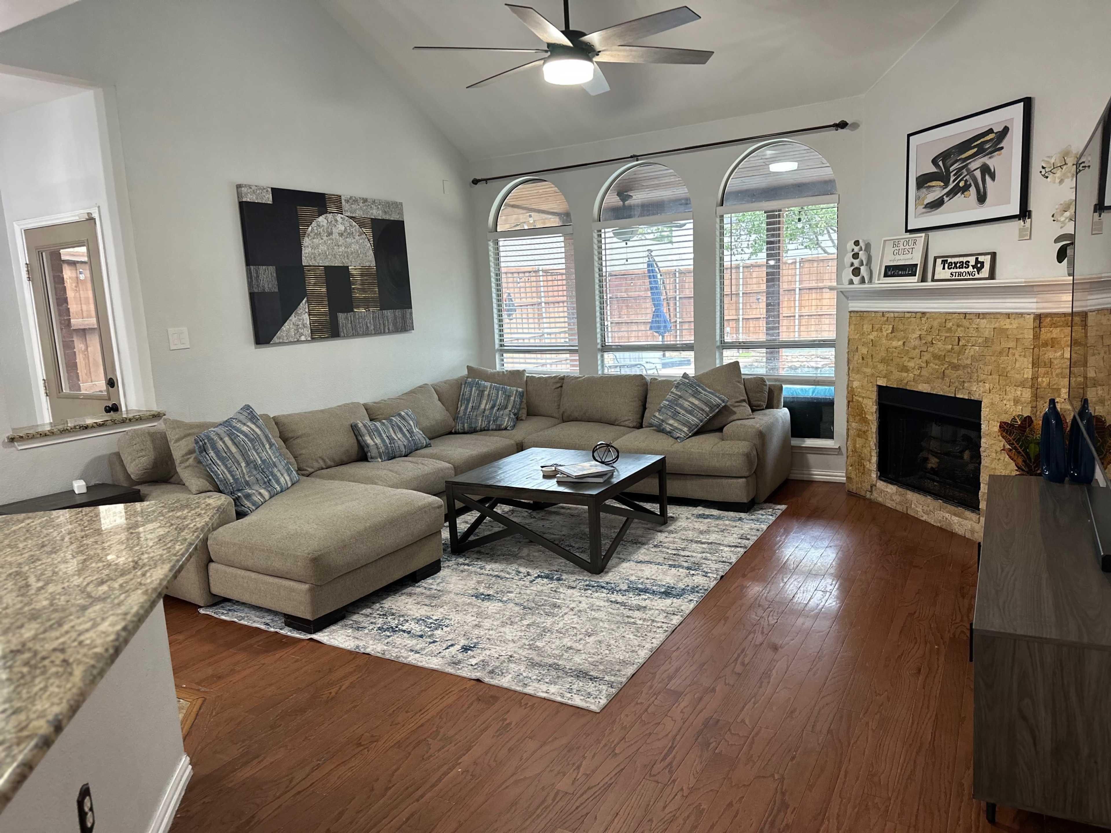 A living room with a large sectional sofa, a coffee table, and a stone fireplace, all under a ceiling fan and natural light from arched windows.
