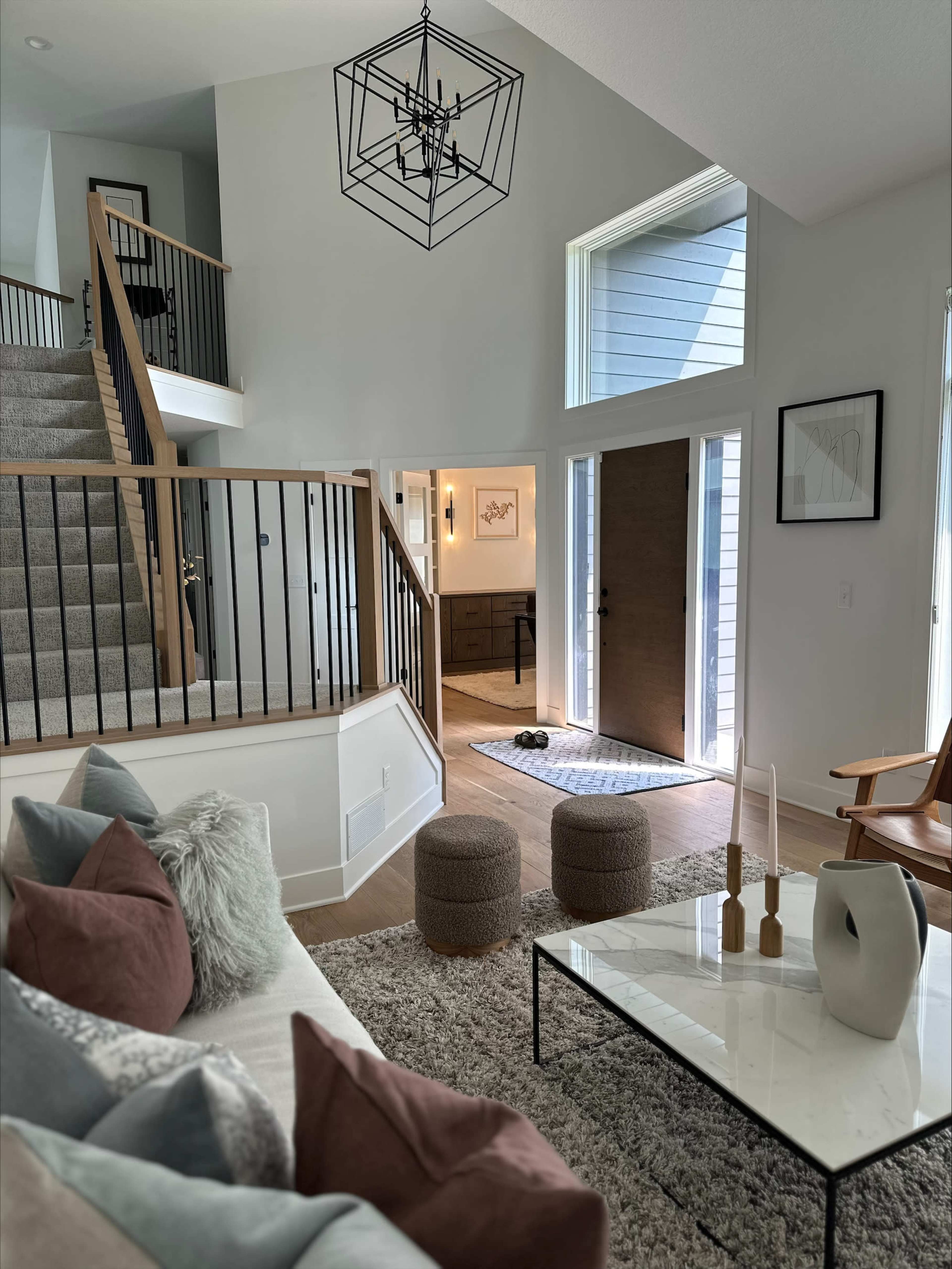 The scene features a modern interior of a home with a staircase, a living area adorned with decorative pillows and a coffee table, and a view through an open doorway to a well-lit entryway.