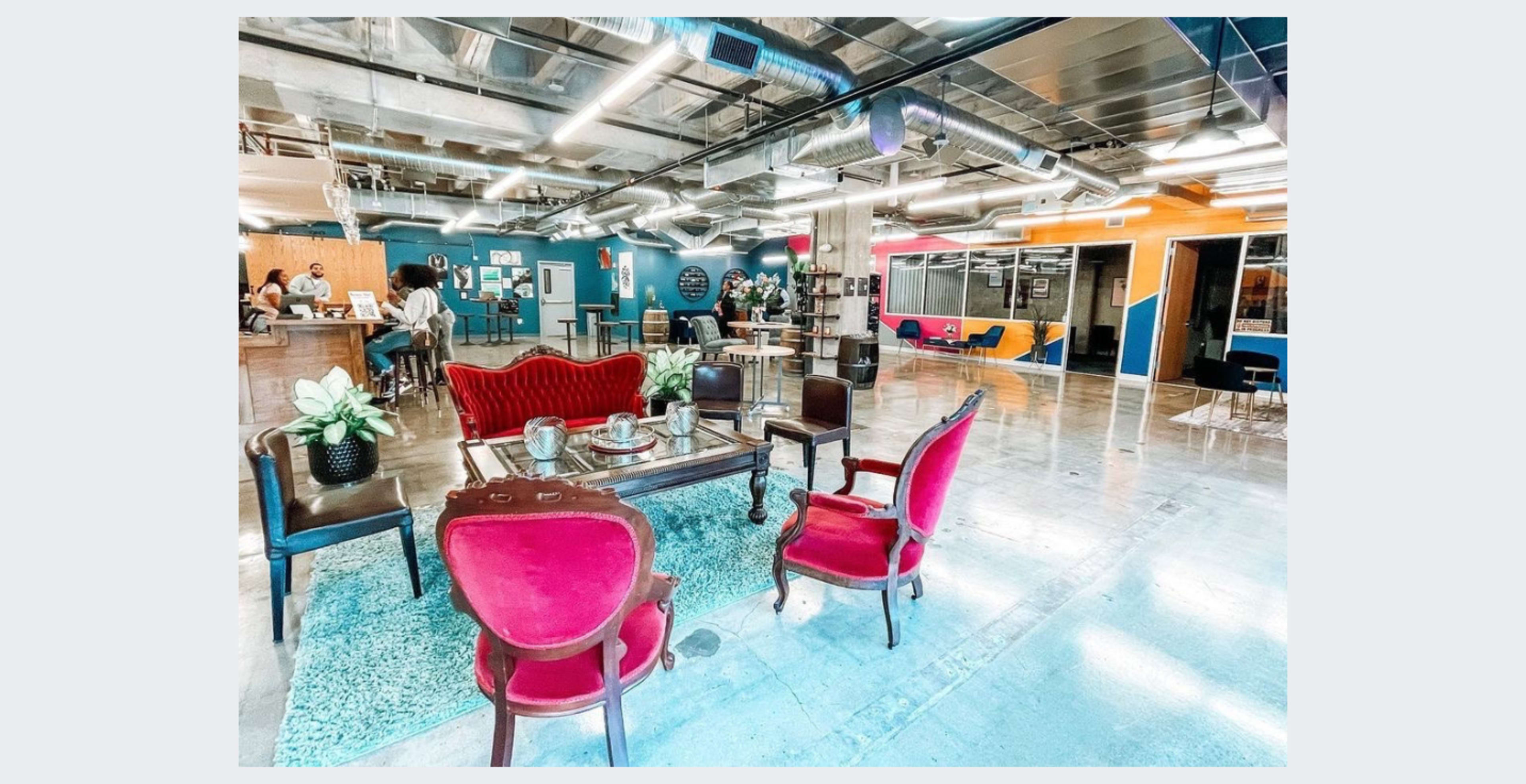 A modern co-working space with a decorative seating area featuring a red velvet sofa and two matching chairs, surrounded by workspace and vibrant walls.