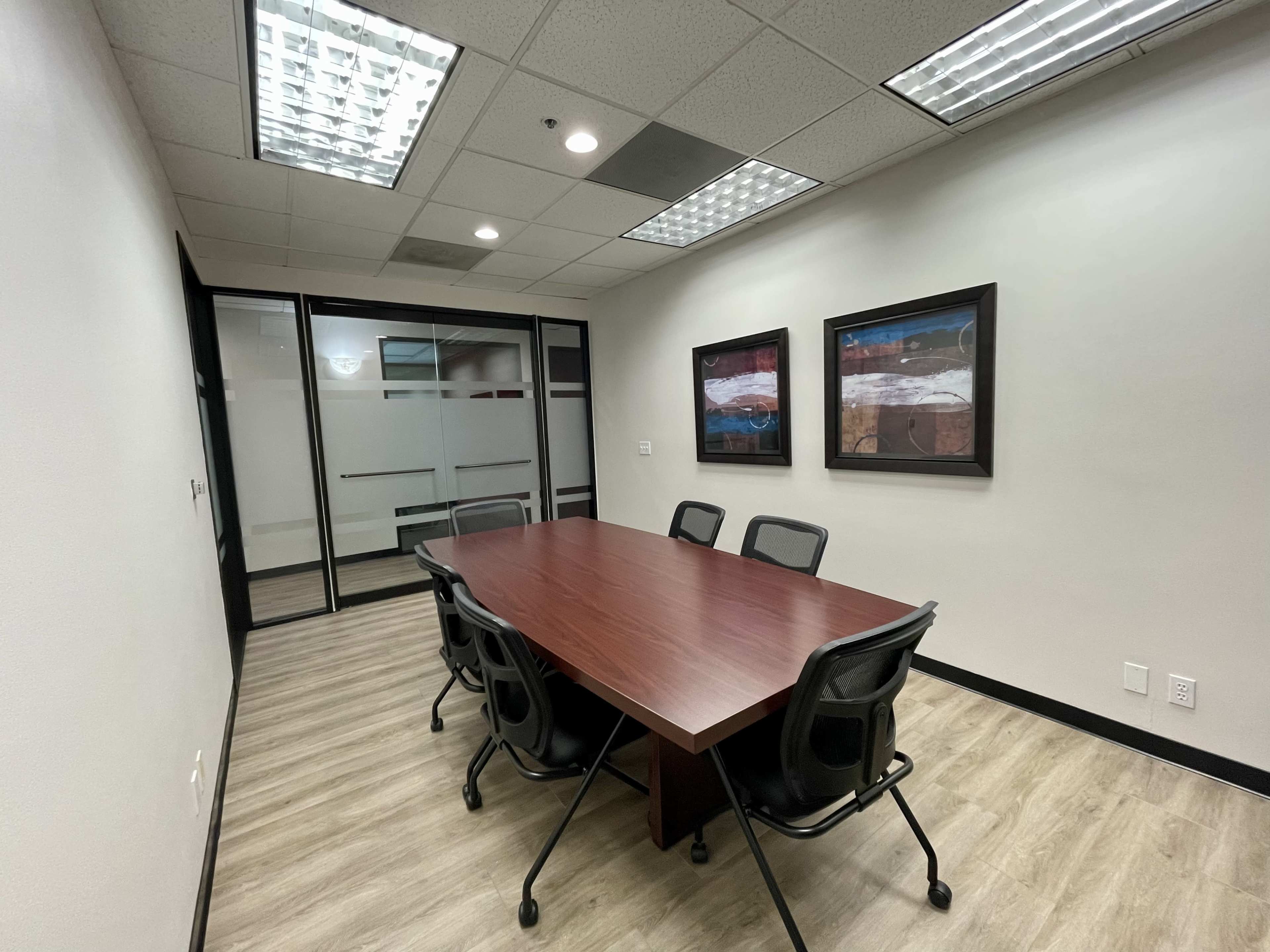 A meeting room features a large wooden table surrounded by six black office chairs, with two framed artworks on the wall and glass partitions.