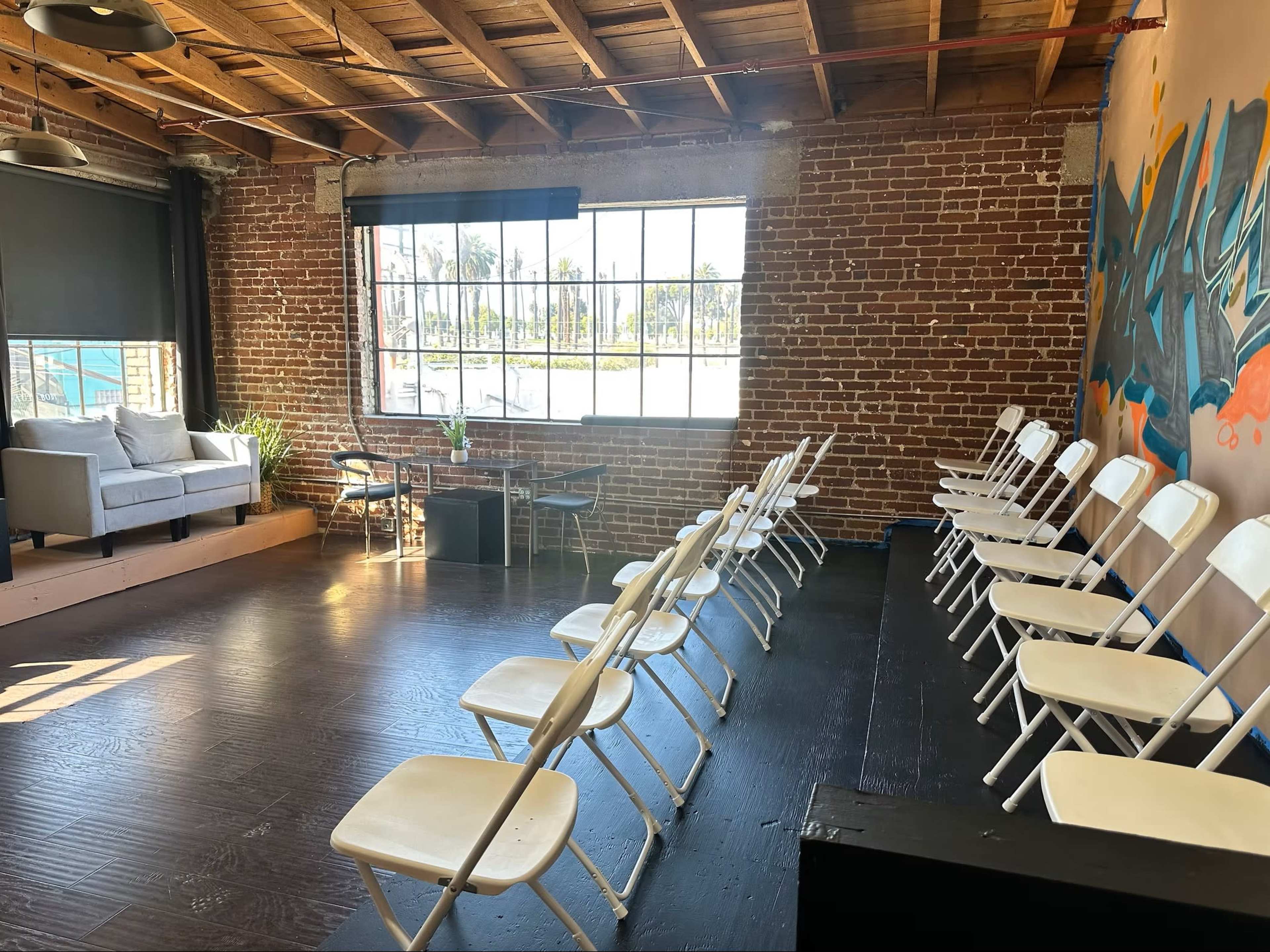 The image shows a spacious room with a row of white chairs arranged facing a small stage, a gray sofa, and large windows showcasing an outdoor view.