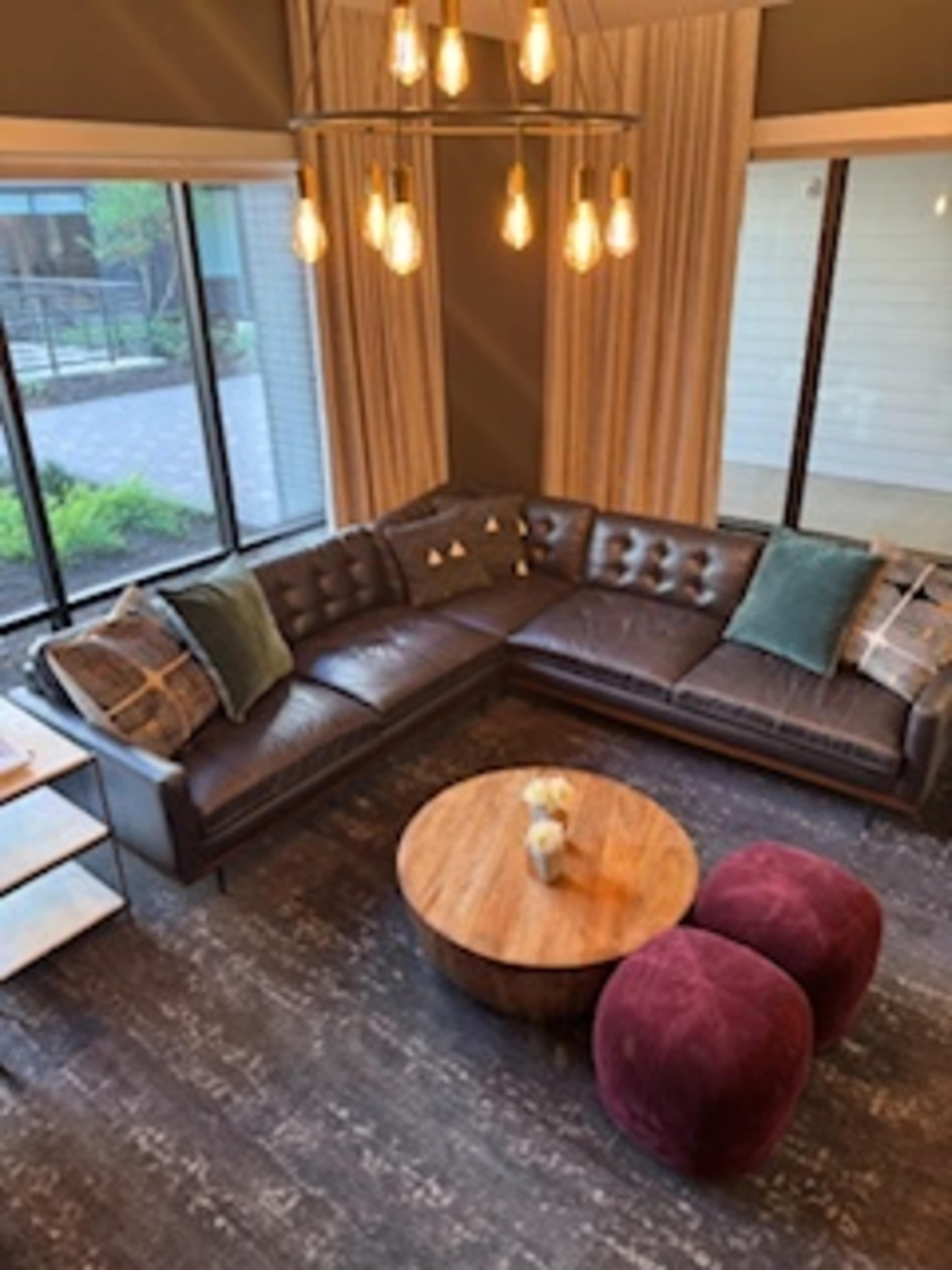 A modern living room features a large leather sectional couch, a round wooden coffee table, and two velvet ottomans, with windows providing natural light.
