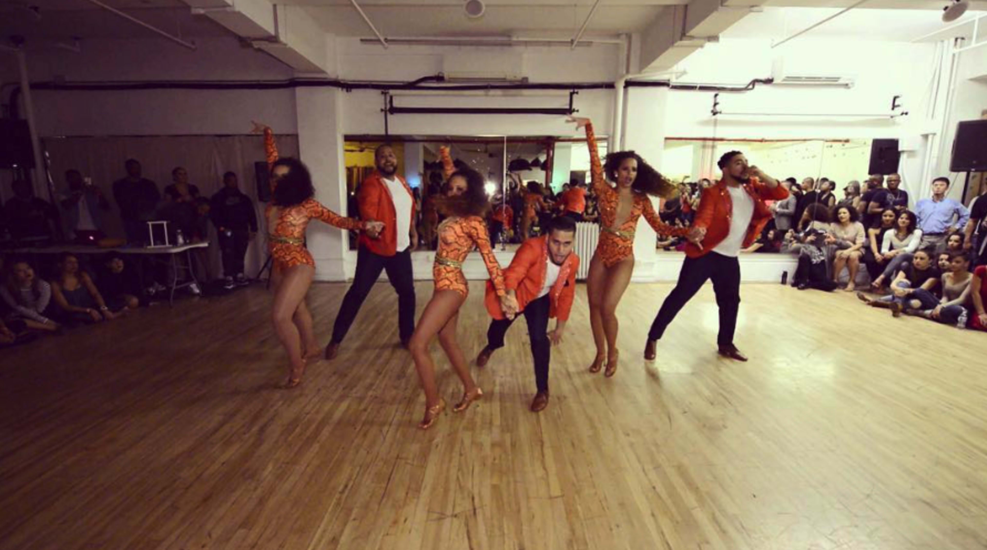 A group of six dancers performs on a wooden floor in a studio, wearing vibrant costumes and showcasing varied poses.