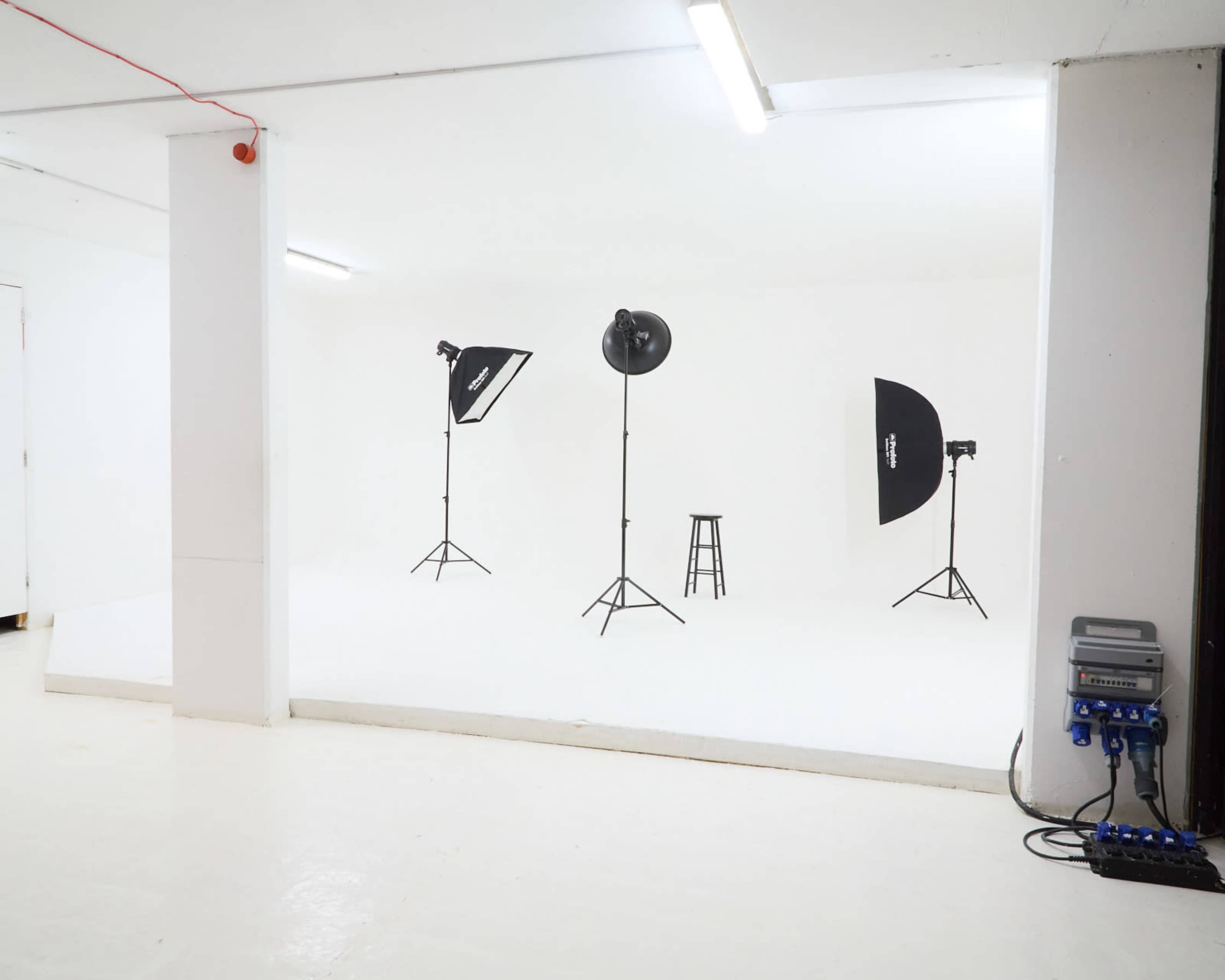 A photography studio with a white backdrop, three light stands, and a stool positioned on a white floor.