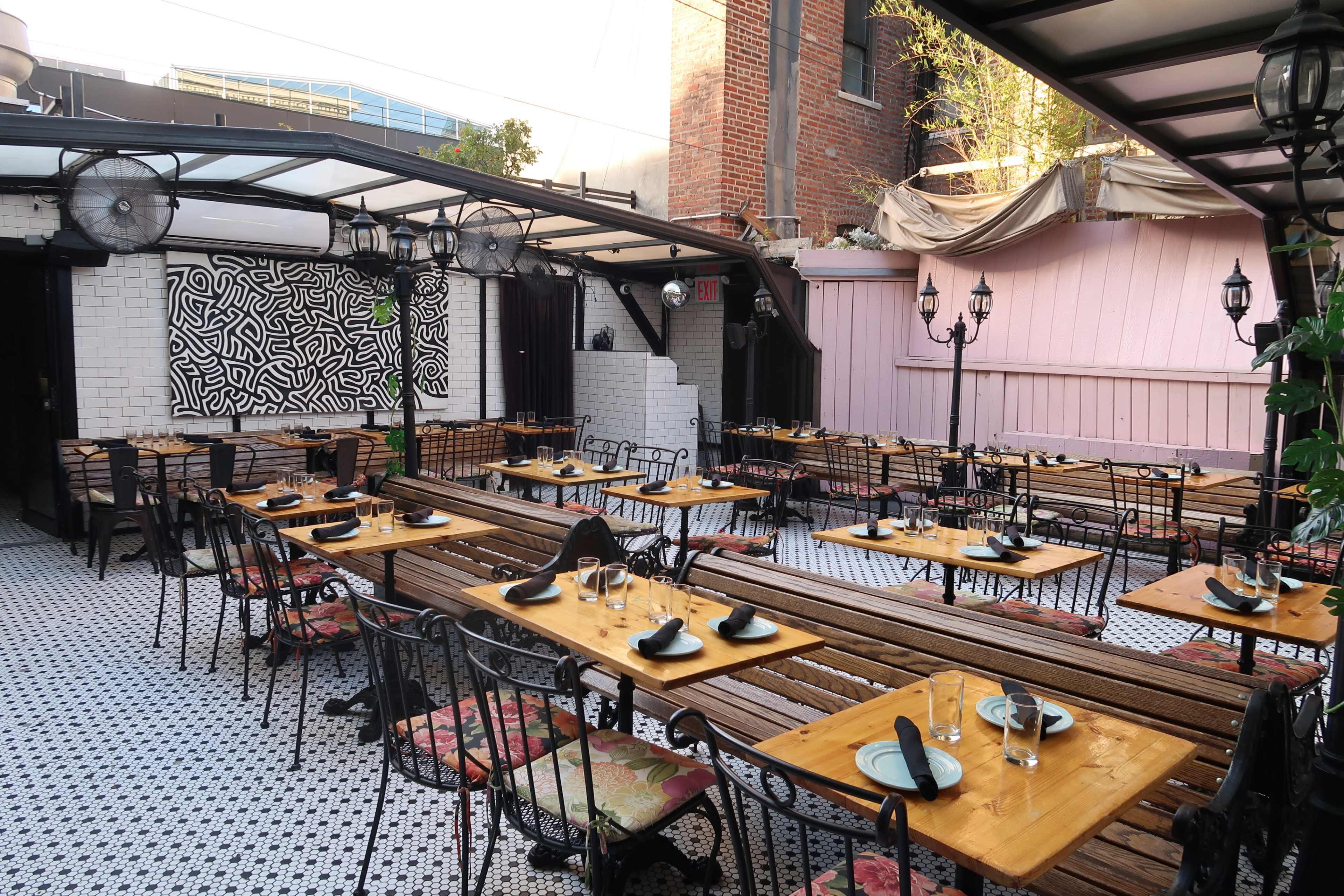 An outdoor restaurant space featuring wooden tables with black plates and glasses, surrounded by brick and painted walls.
