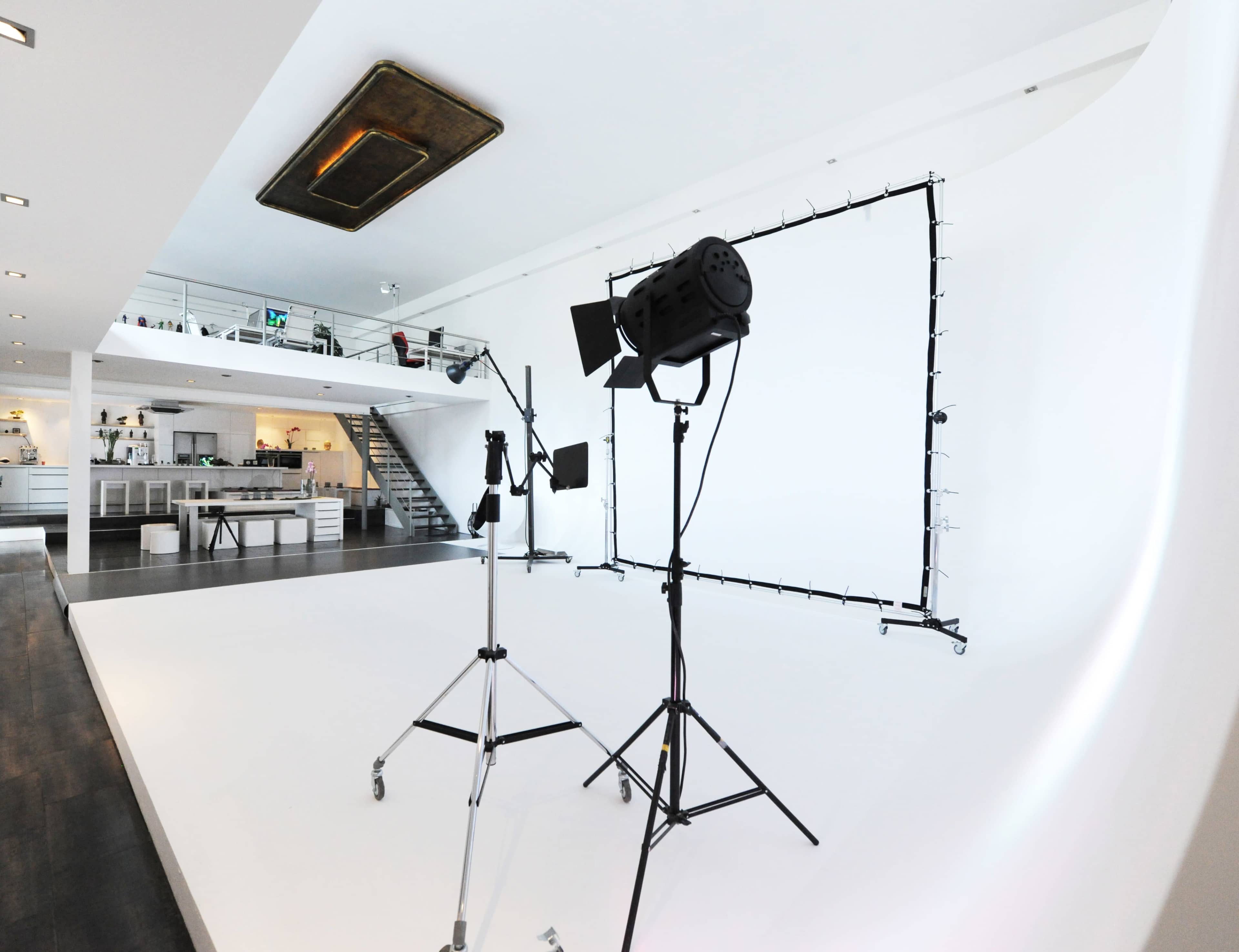 The image shows a photography studio with a white backdrop, lighting equipment set up on stands, and a modern open space featuring a visible staircase and kitchen area in the background.