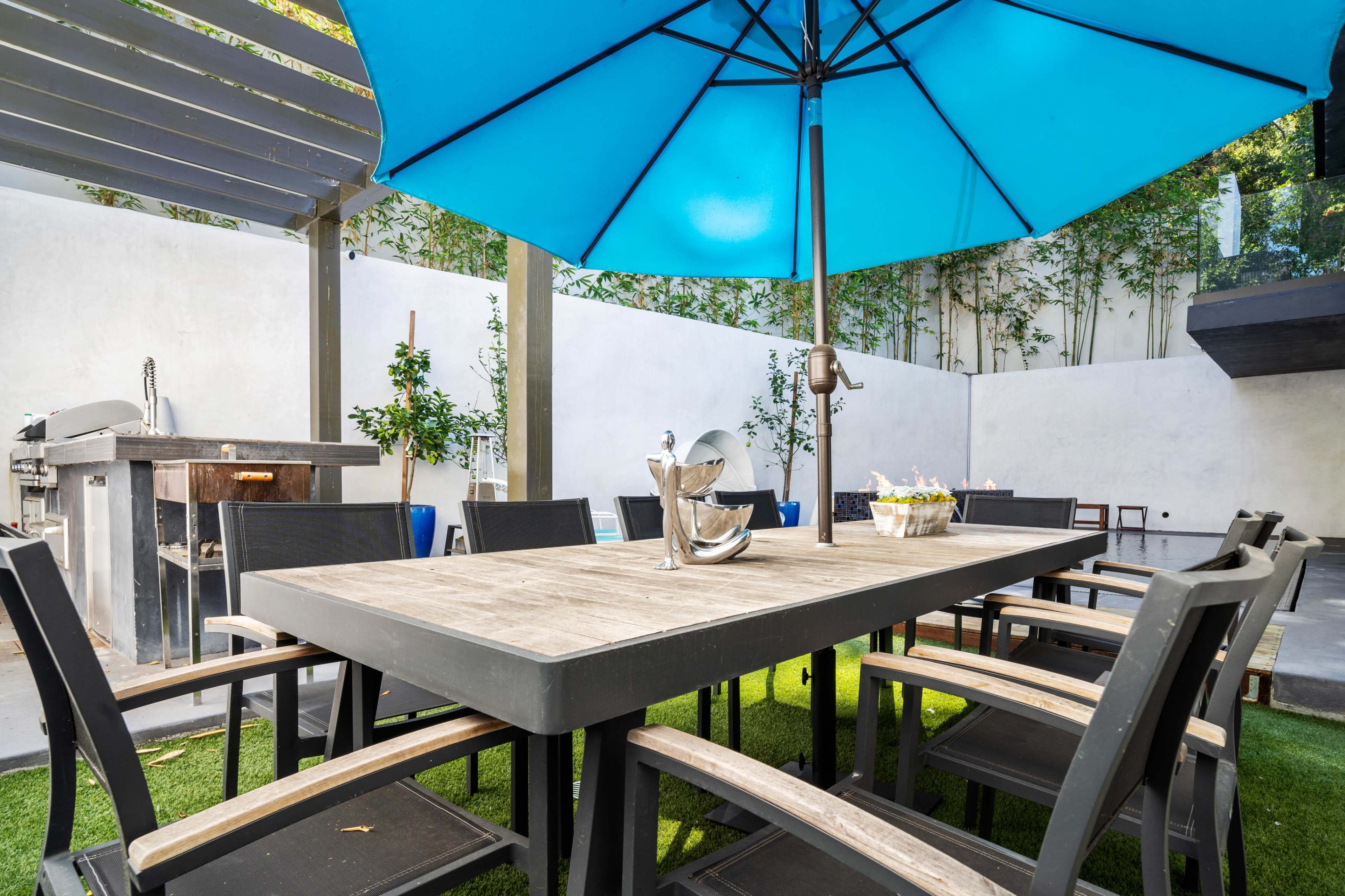 A large outdoor dining table is surrounded by black chairs under a blue umbrella in a modern patio setting.