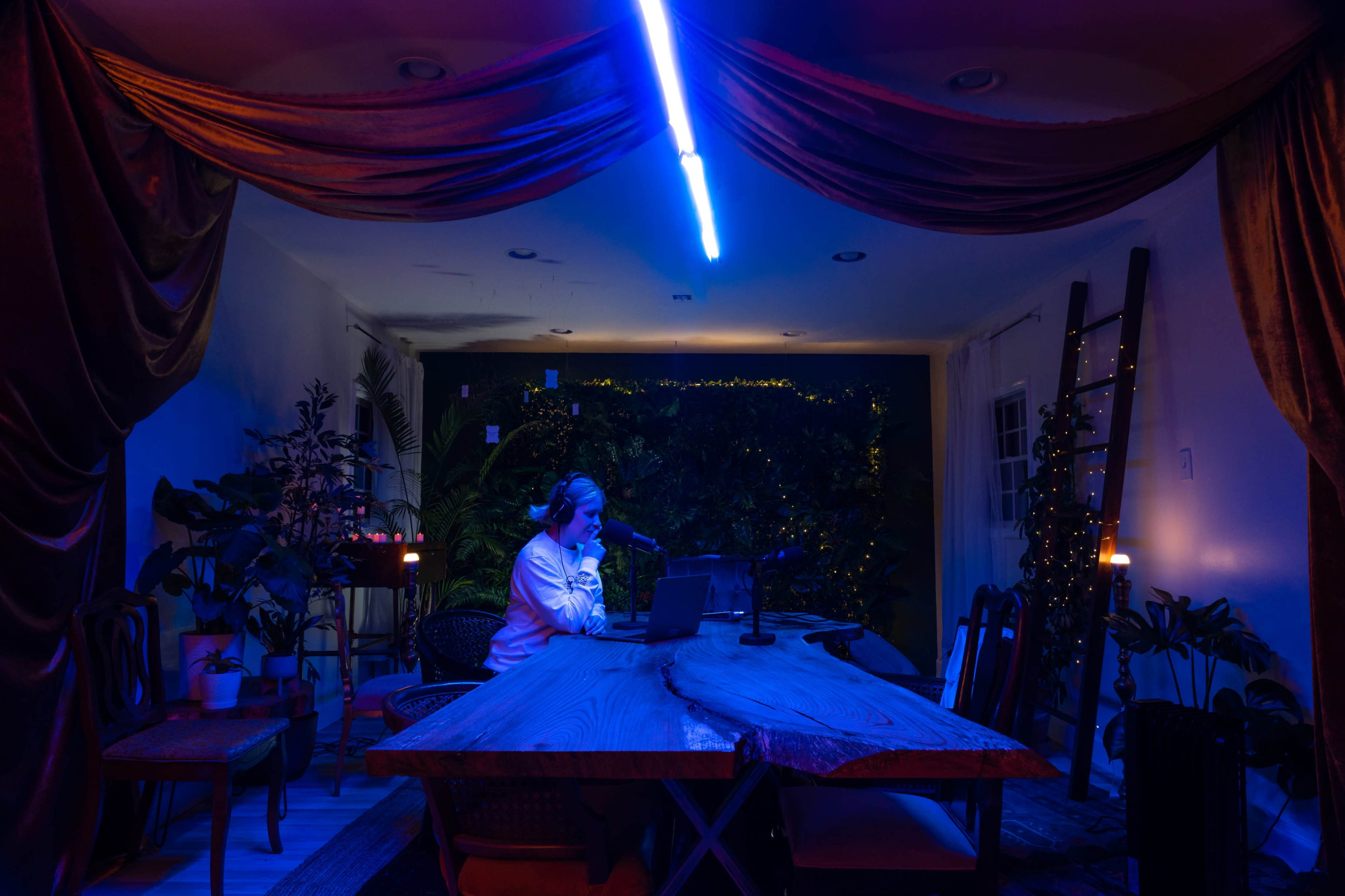 A person sits at a wooden table in a dimly lit room decorated with plants and draped fabric, using a microphone while working on a laptop.