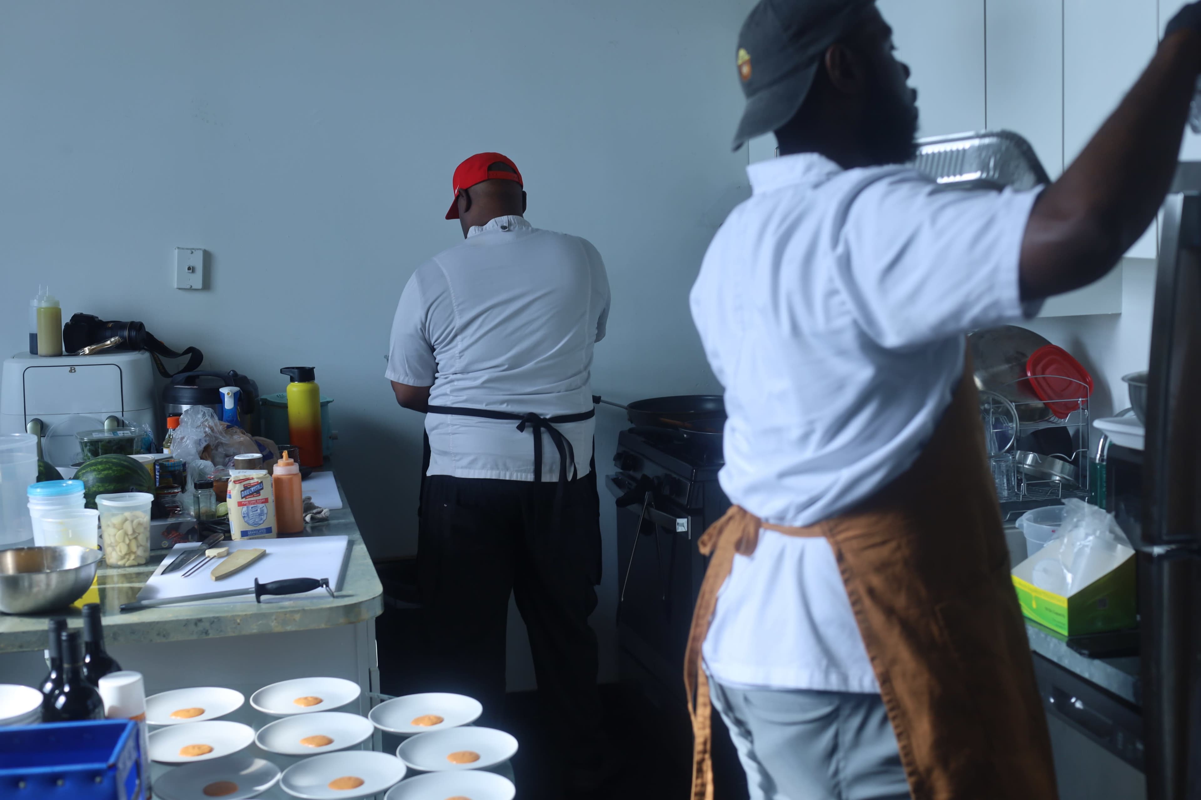 Two cooks are preparing food in a kitchen, with one focused on the stove and the other arranging ingredients on a table.