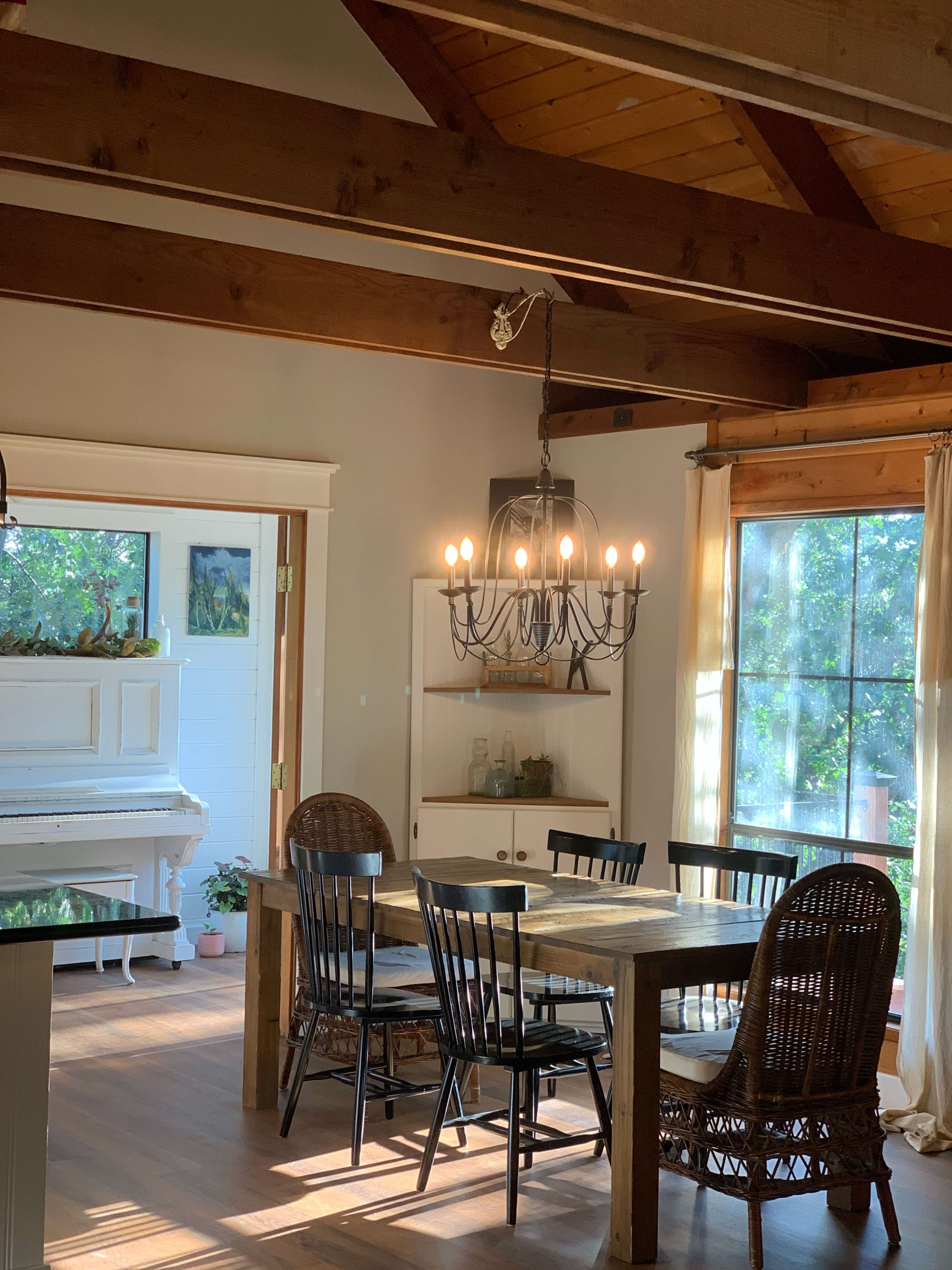 A dining area features a wooden table surrounded by black chairs and a chandelier hanging from a timber ceiling, with a white piano and large windows in the background.