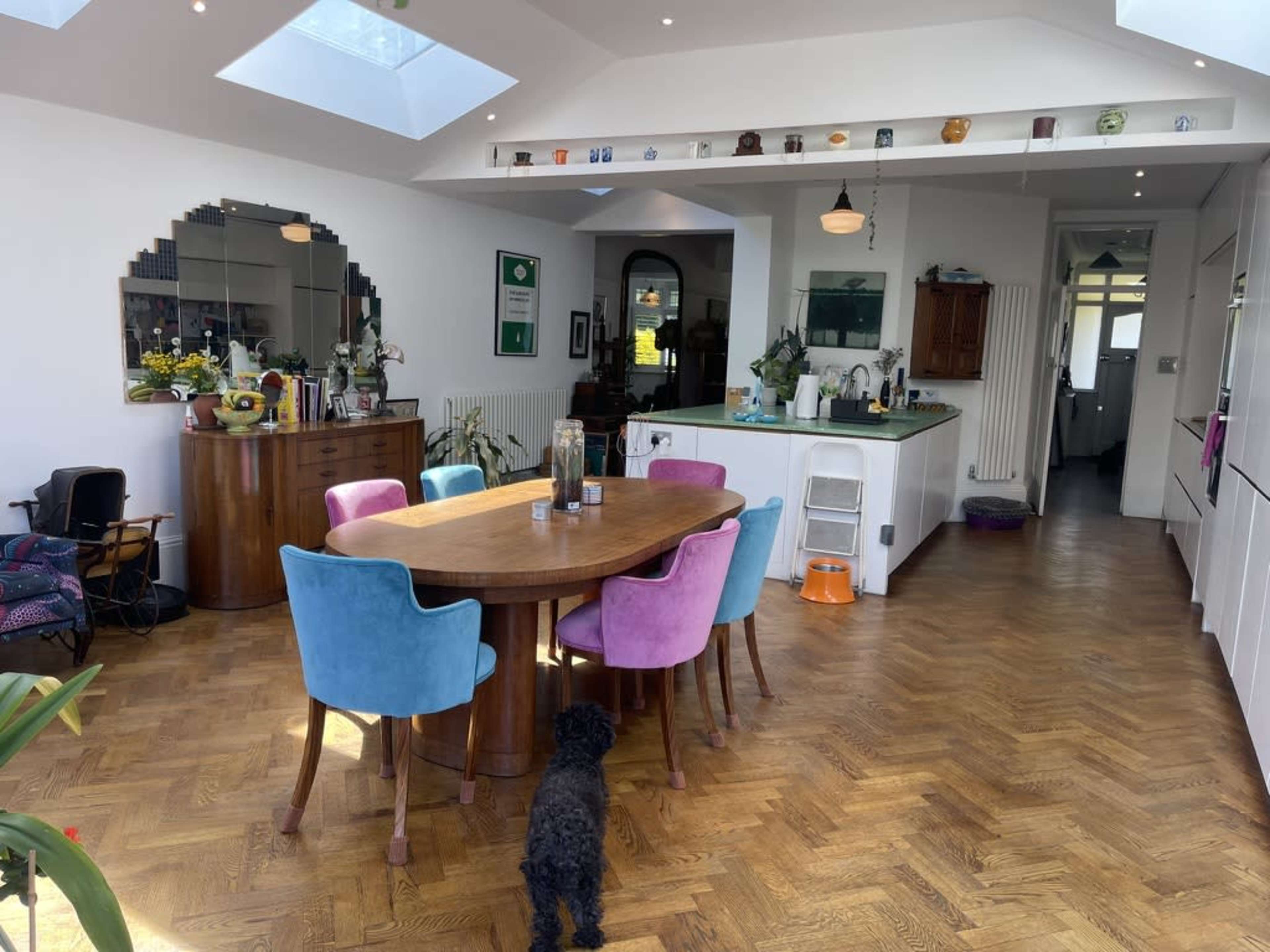 A dining area features a large wooden table surrounded by colorful chairs, with a kitchen visible in the background.