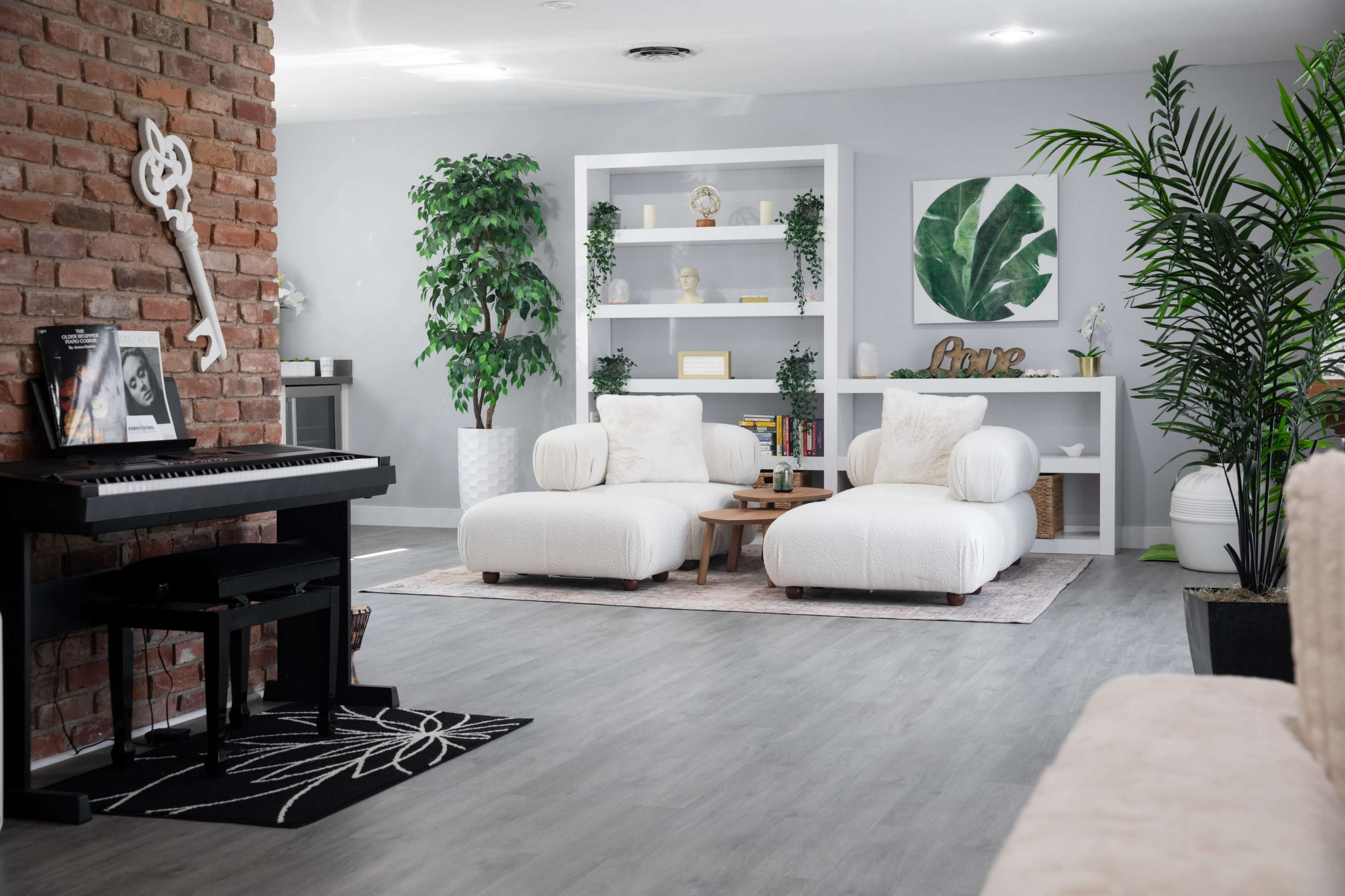 The image shows a modern living room with a piano, two white armchairs, a coffee table, and decorative shelves against a brick wall.
