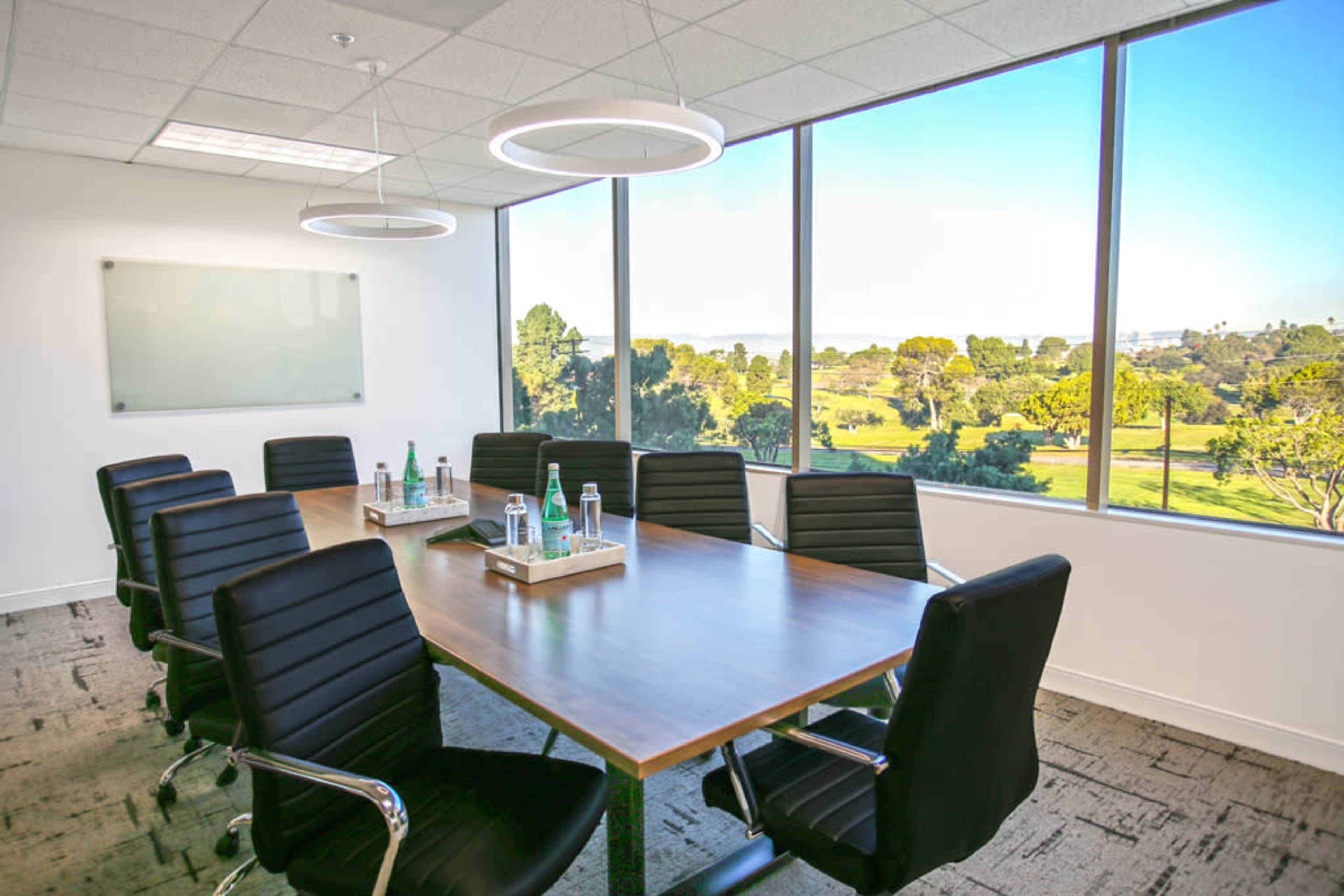 A modern conference room features a large wooden table surrounded by black chairs, with large windows displaying a view of green outdoor landscape.