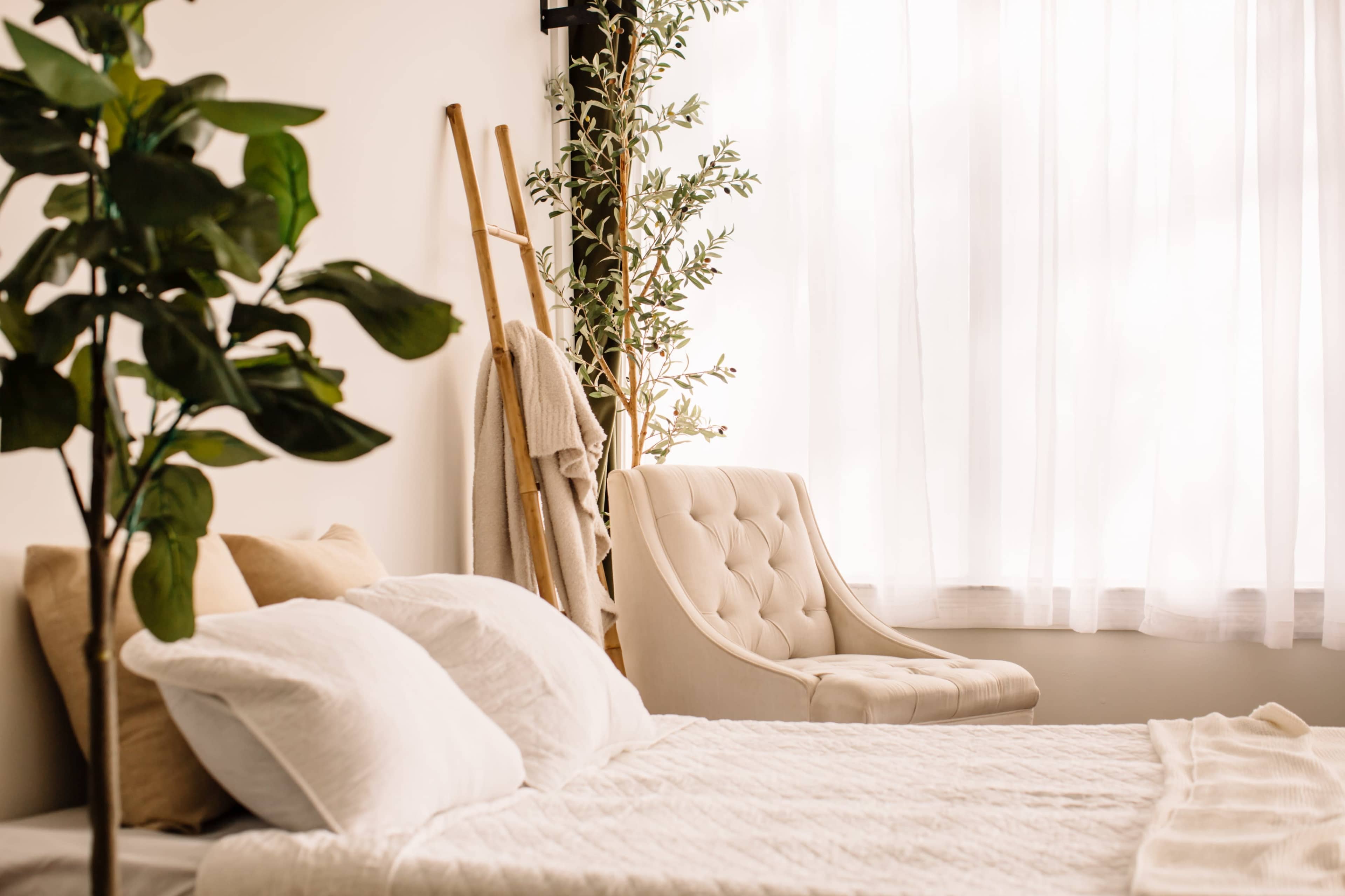 A bedroom features a white bed with pillows, a cream-colored chair, and a plant beside a large window draped with sheer curtains.
