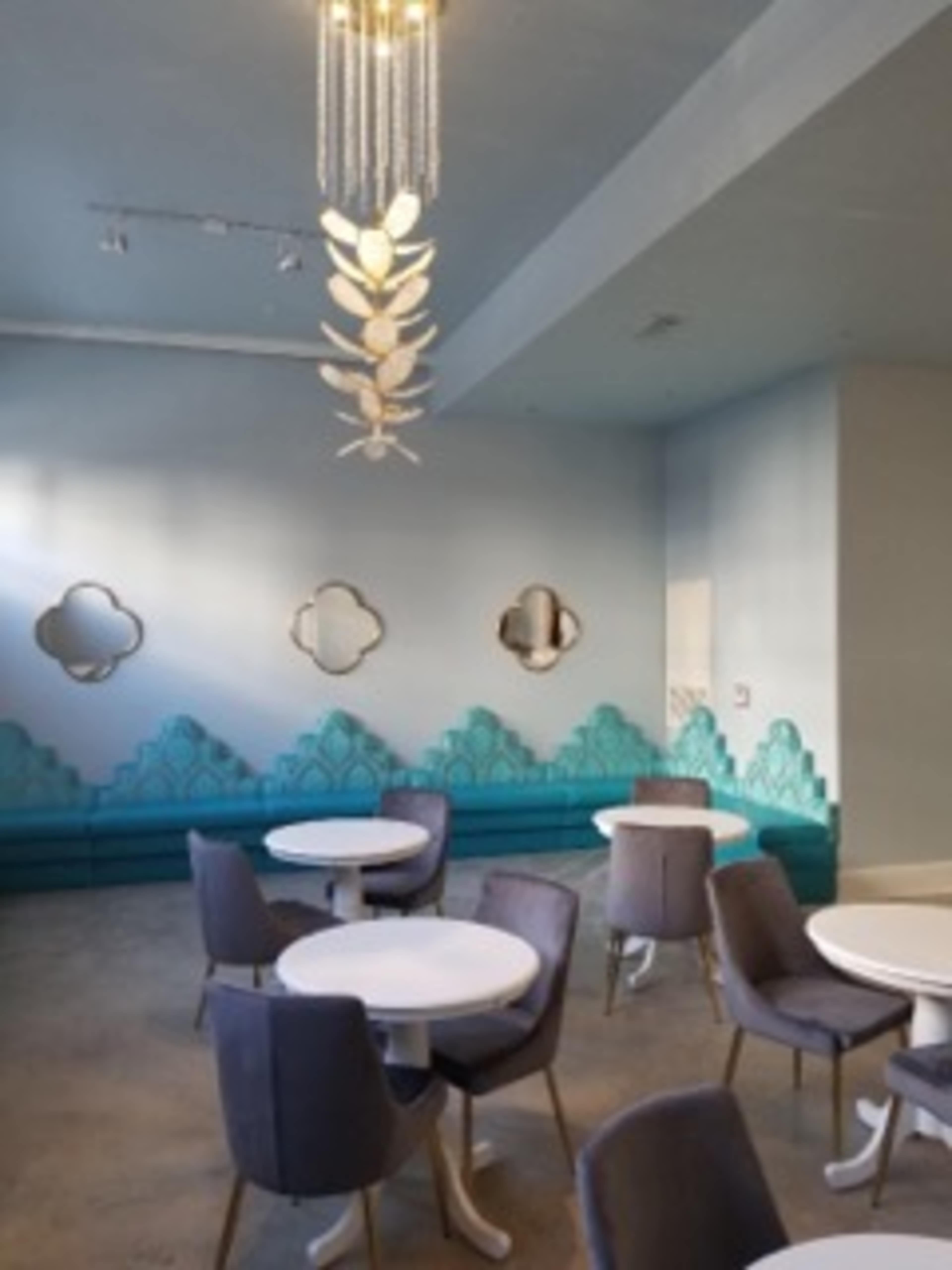 The image shows a modern café interior with circular tables, gray chairs, a teal decorative wall, and a unique chandelier.