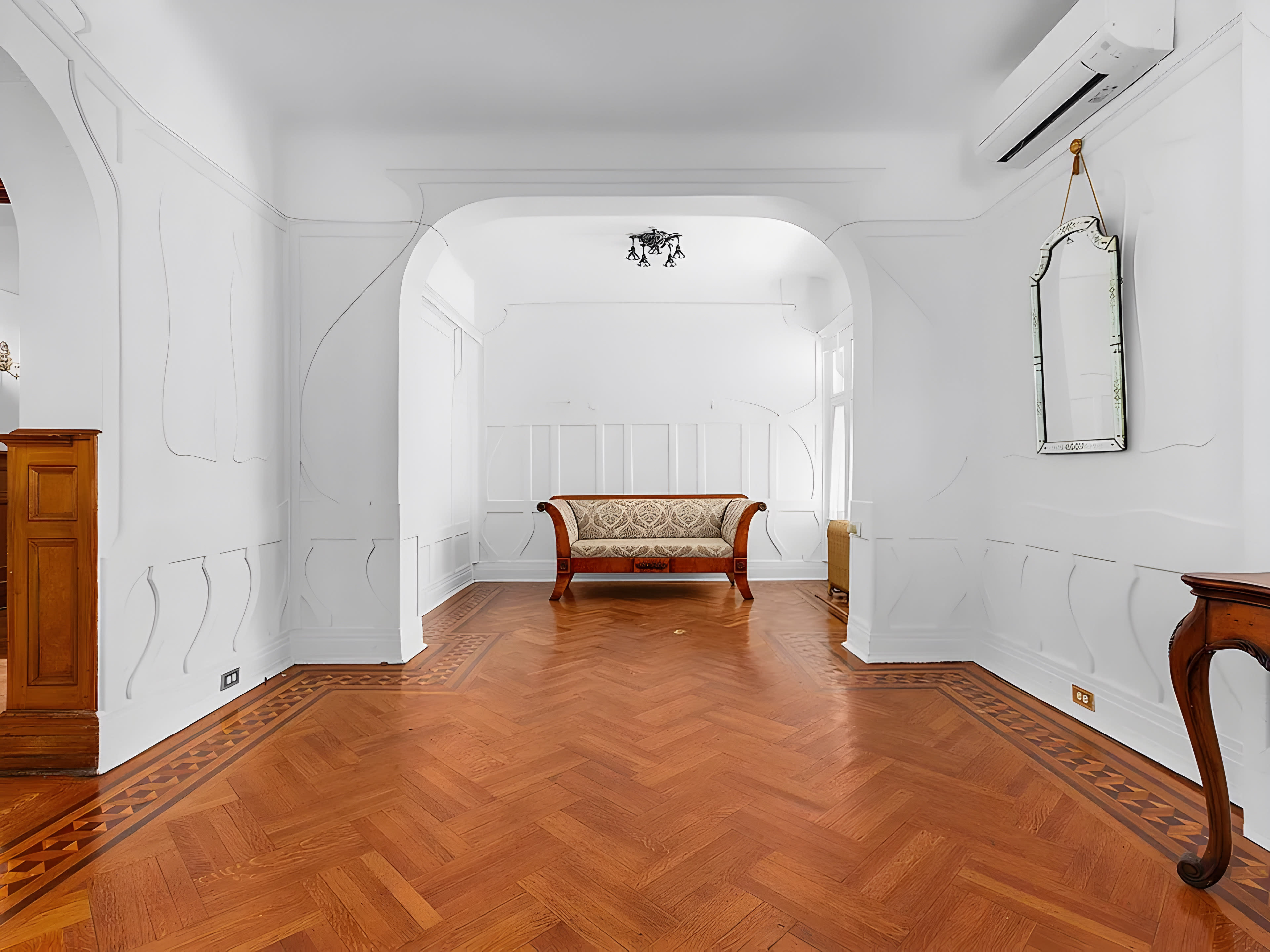 A bright, empty room featuring a wooden floor with a herringbone pattern, a decorative couch in the center, and an ornate mirror on the wall.