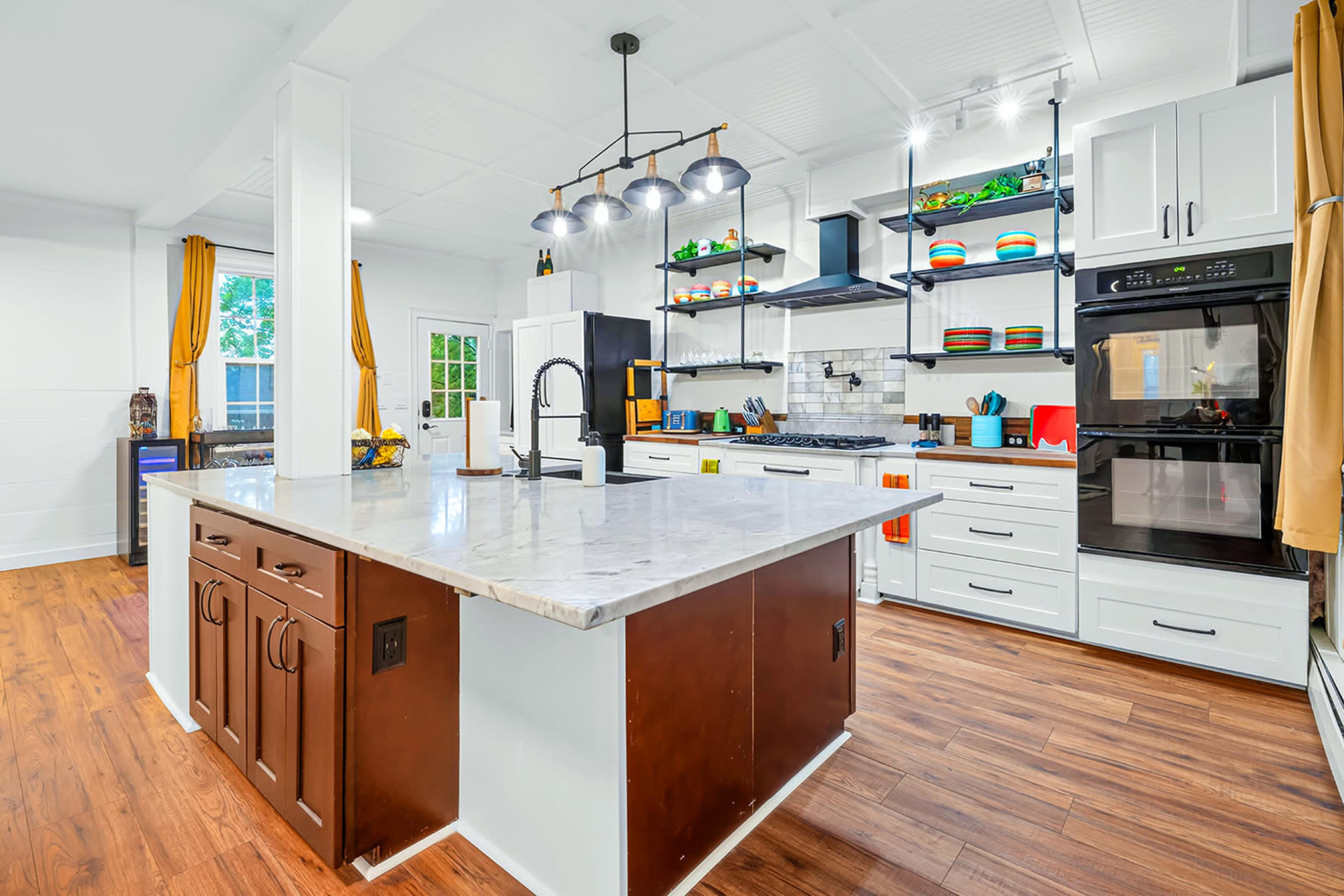 The image shows a modern kitchen featuring white cabinetry, a marble island with dark wood accents, and an arrangement of colorful dishware on open shelves.