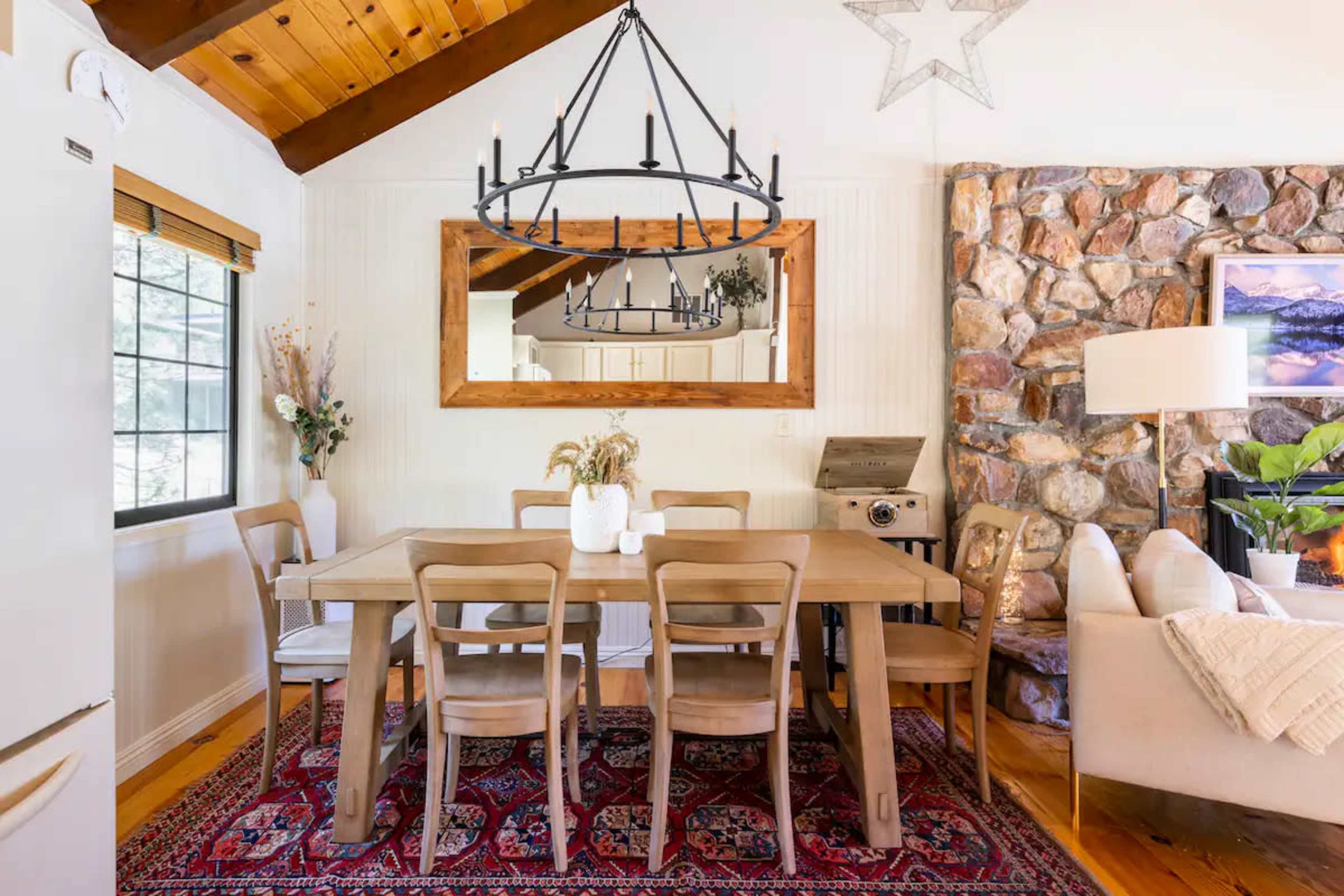A wooden dining table is set in a bright room with a stone wall, large mirror, and a chandelier overhead.