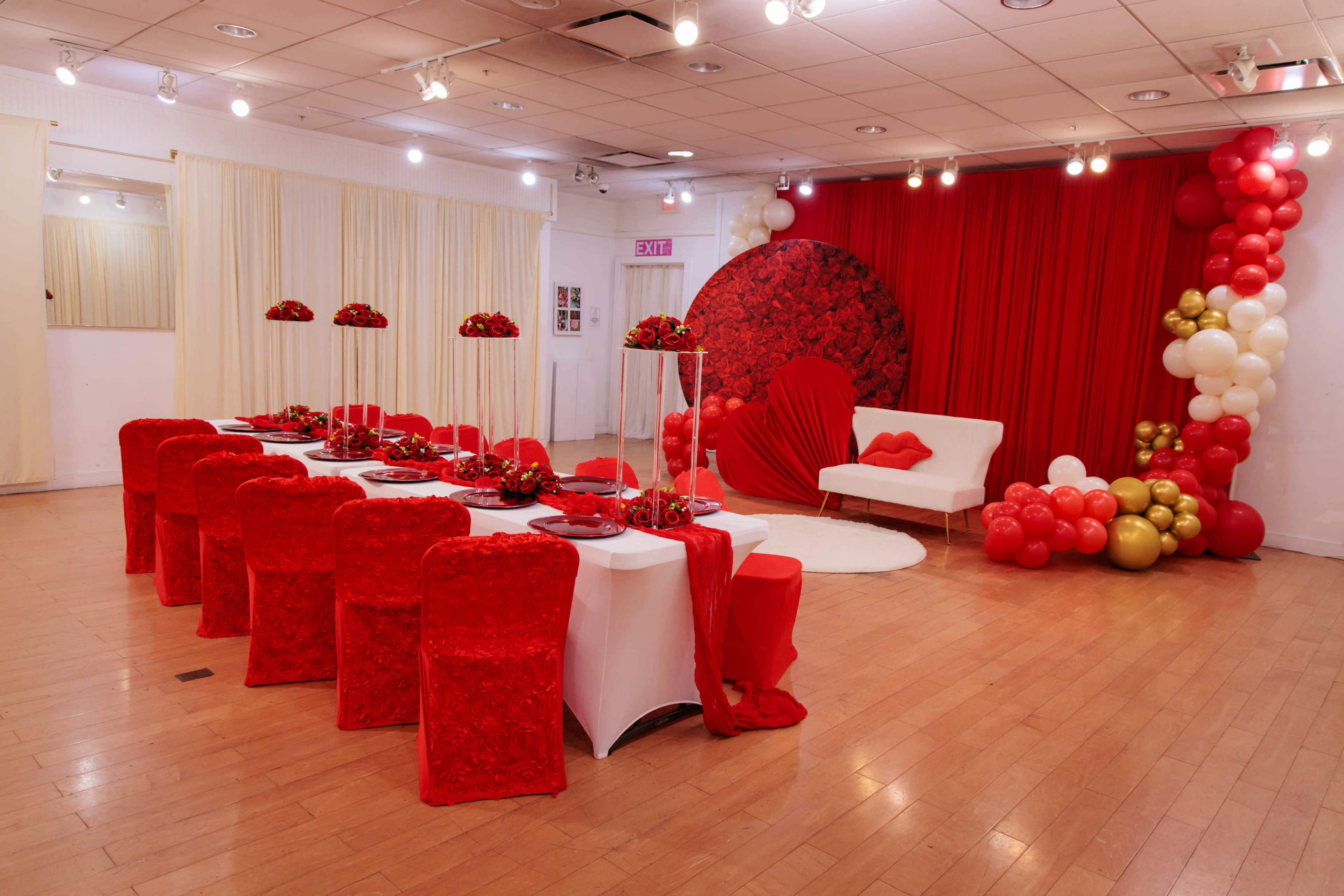 The image shows a decorated event space with a long table set in red and white, surrounded by red chairs, and featuring floral arrangements and a backdrop with red drapery and balloon decor.