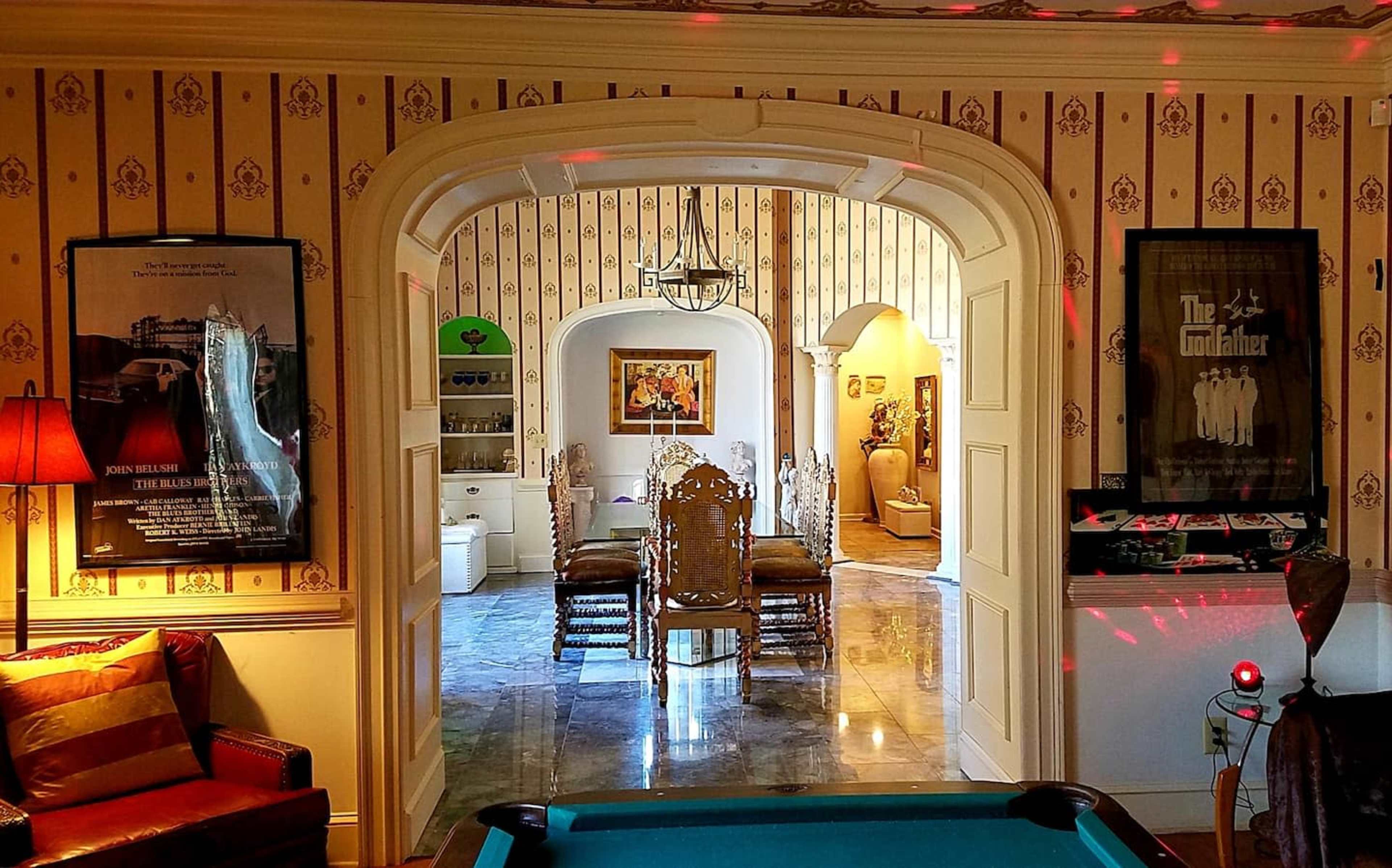 The image shows an interior space with a pool table in the foreground, an arched doorway leading to a dining area, and walls adorned with patterned wallpaper and framed movie posters.