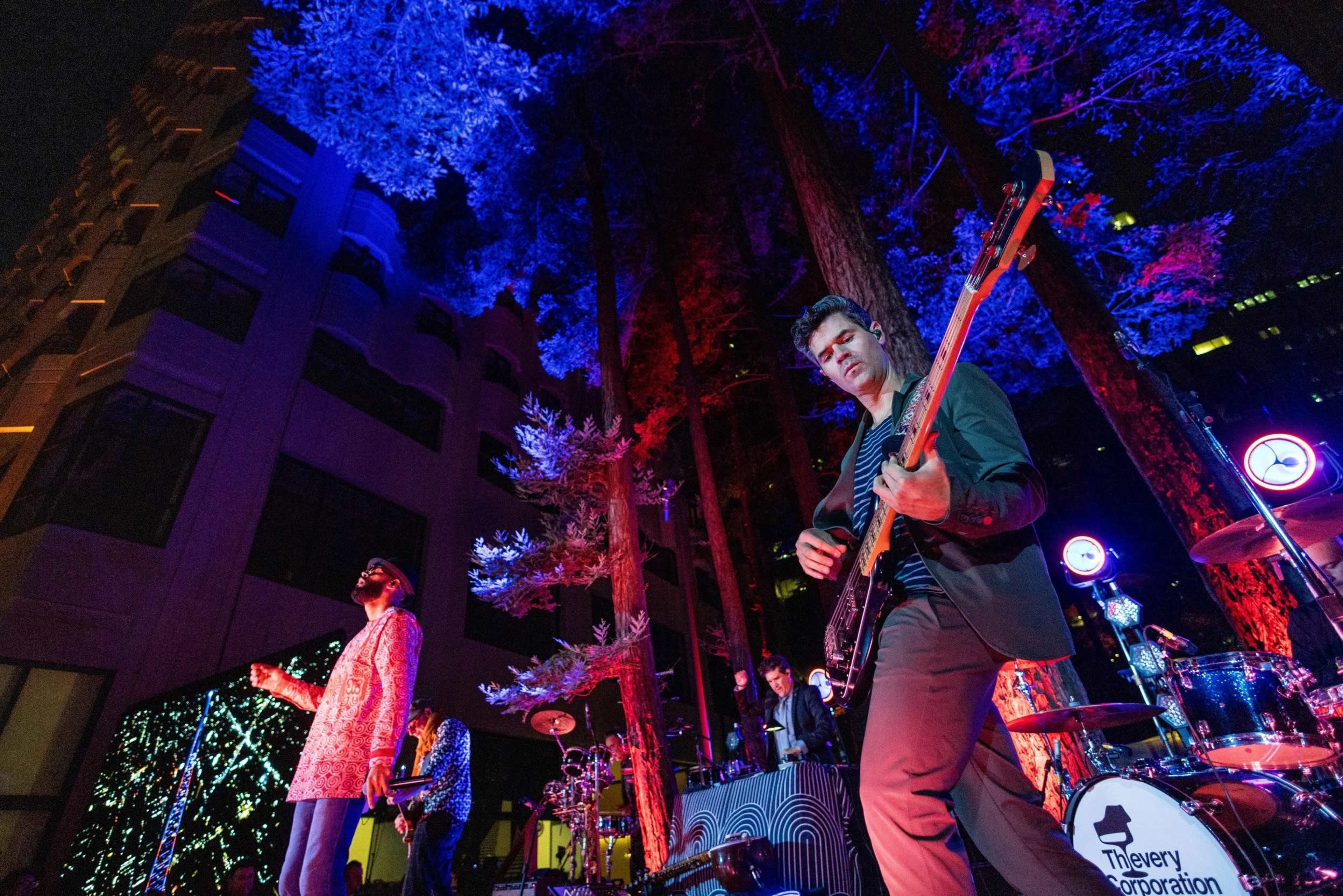 A band performs on an outdoor stage surrounded by tall trees illuminated in vibrant colors at night.