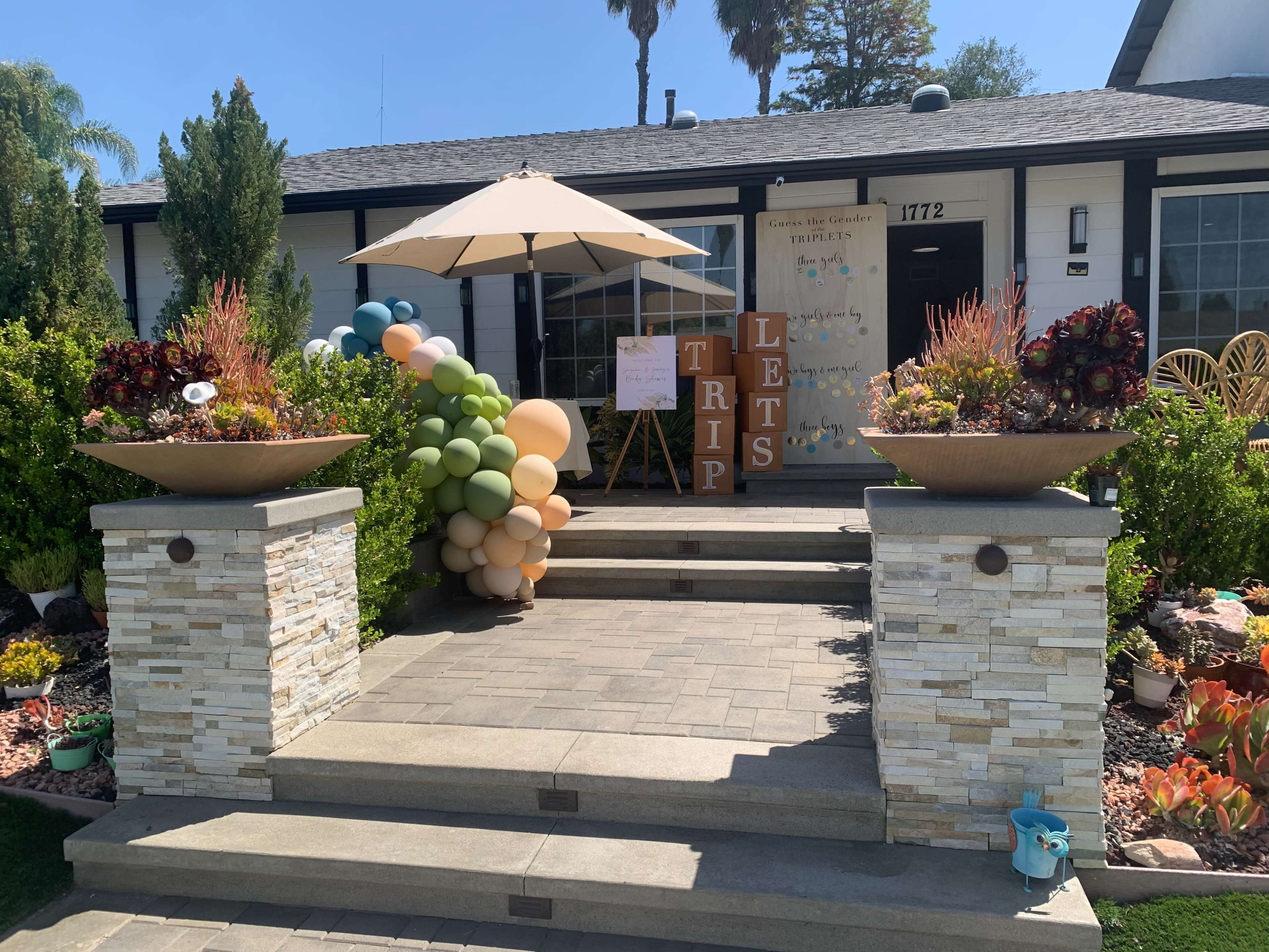 A residential entrance features a well-maintained front yard with decorative planters, a balloon arch, and a sign promoting a trip.