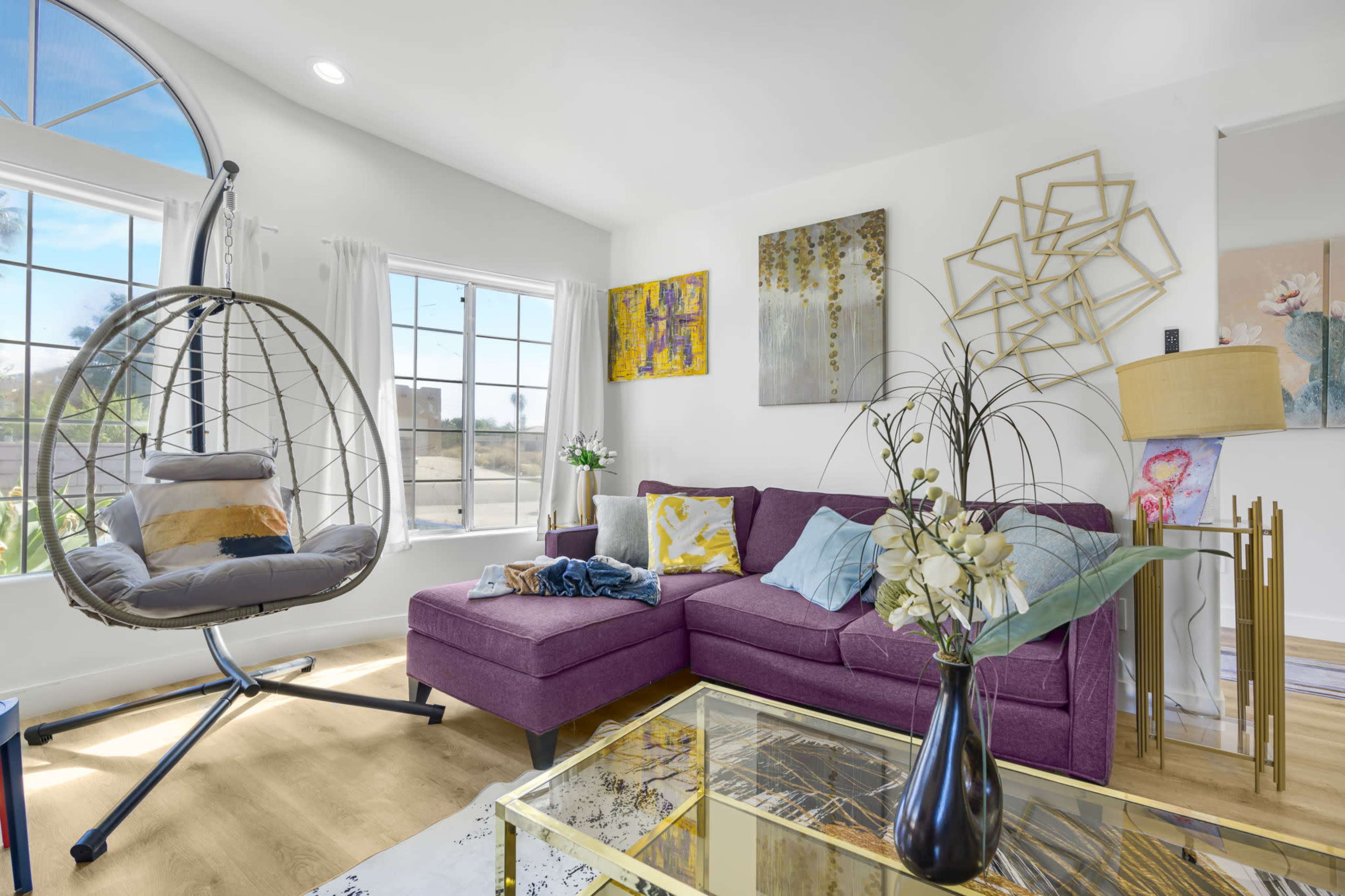 The living room features a purple sectional sofa, a hanging egg chair, a coffee table, and large windows that let in natural light.