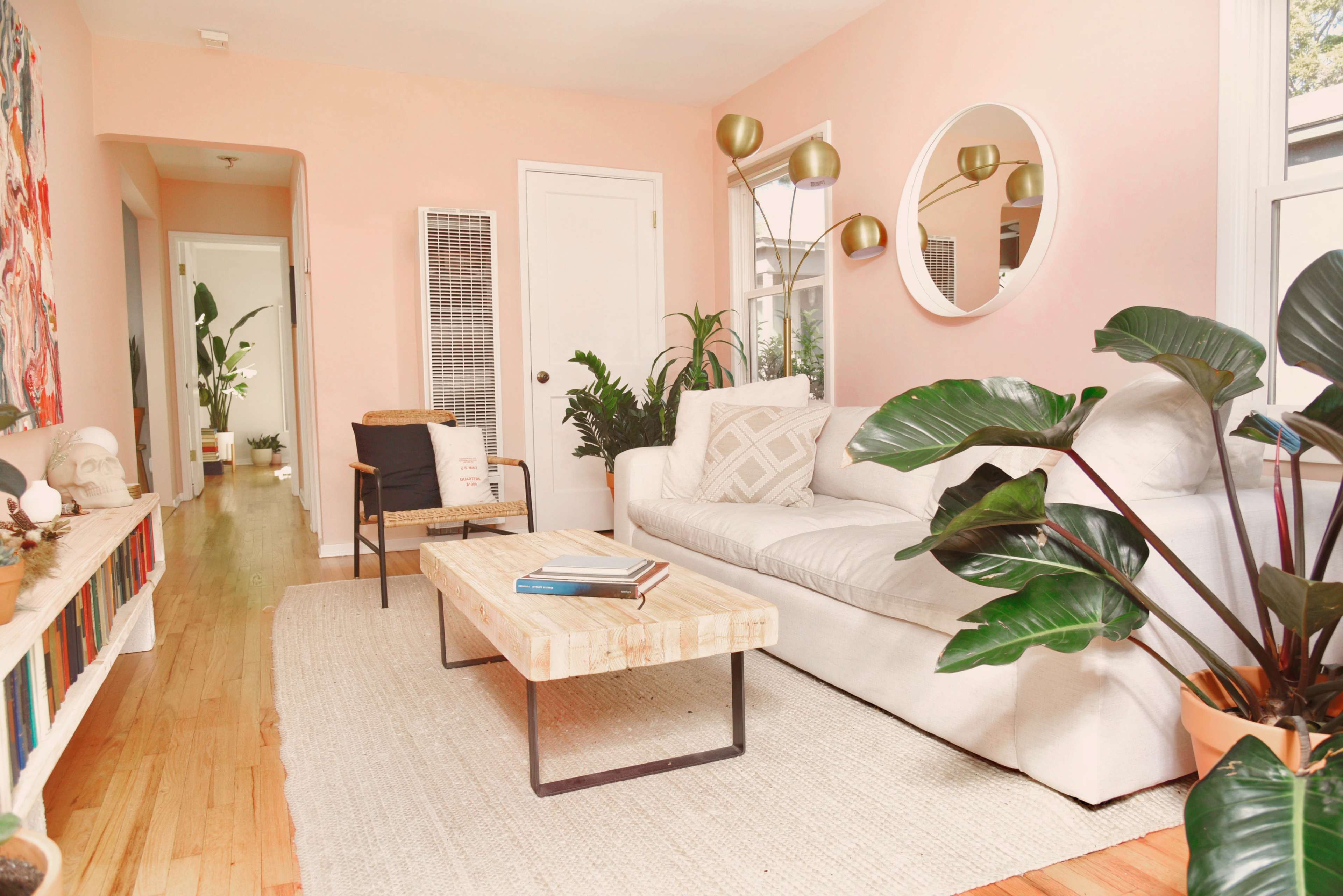 A light-filled living room with a white sofa, a wooden coffee table, and plants arranged near a pink wall, leading to a hallway with additional greenery.