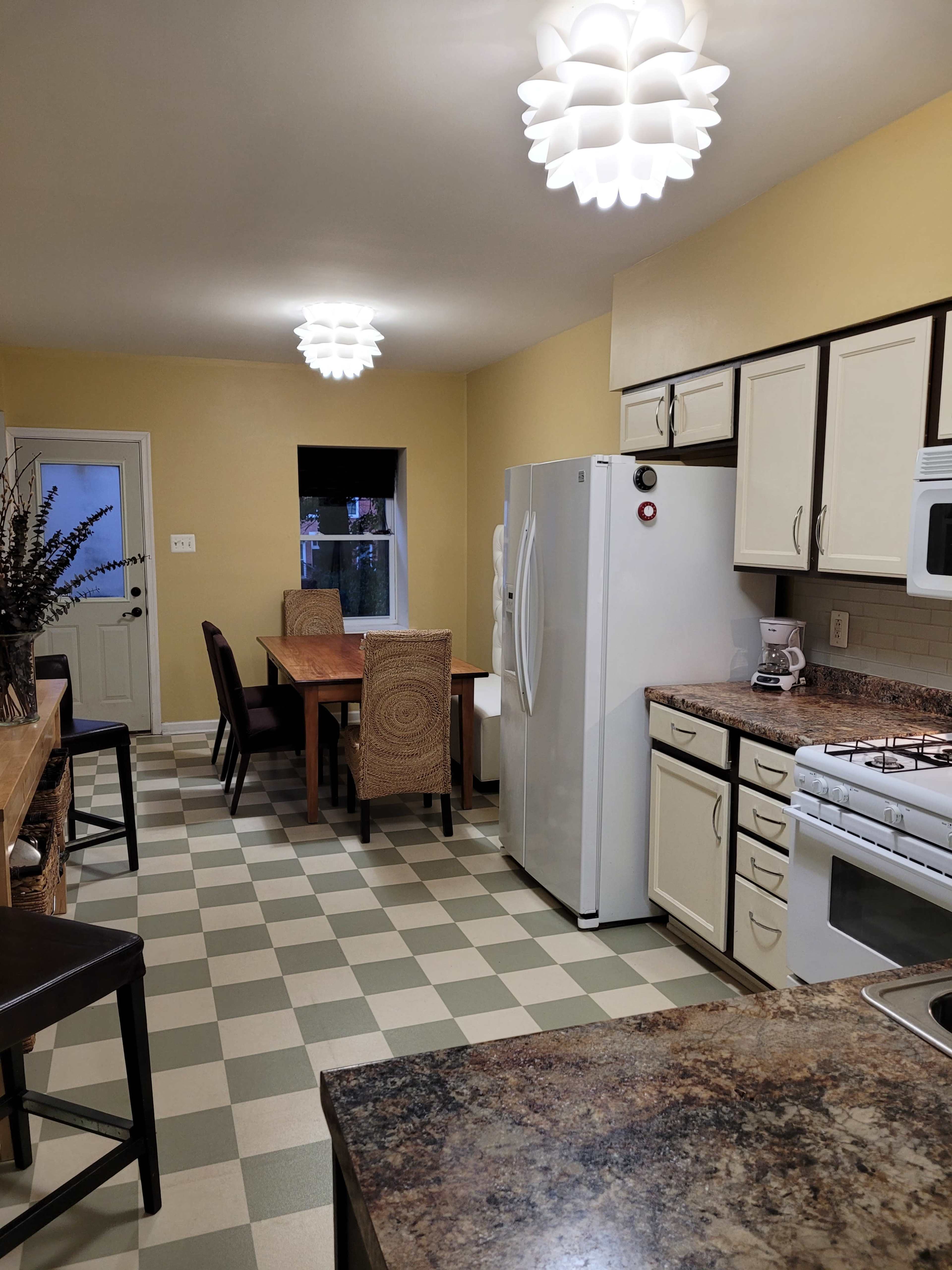 A kitchen features a dining table with chairs, a white refrigerator, and a stove, all illuminated by modern pendant lights.