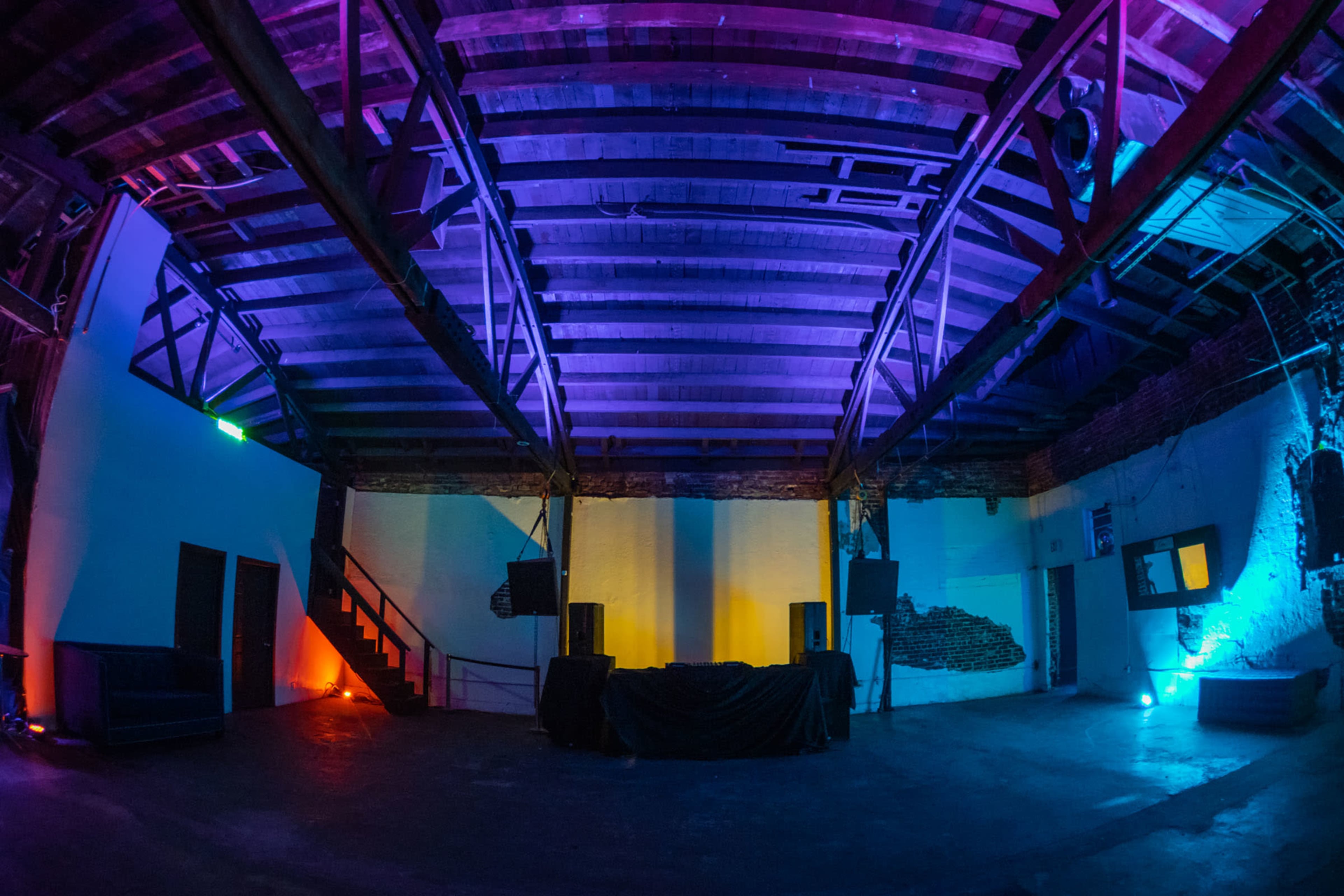 The image shows an empty, brightly lit event space with colored lighting and exposed wooden beams.