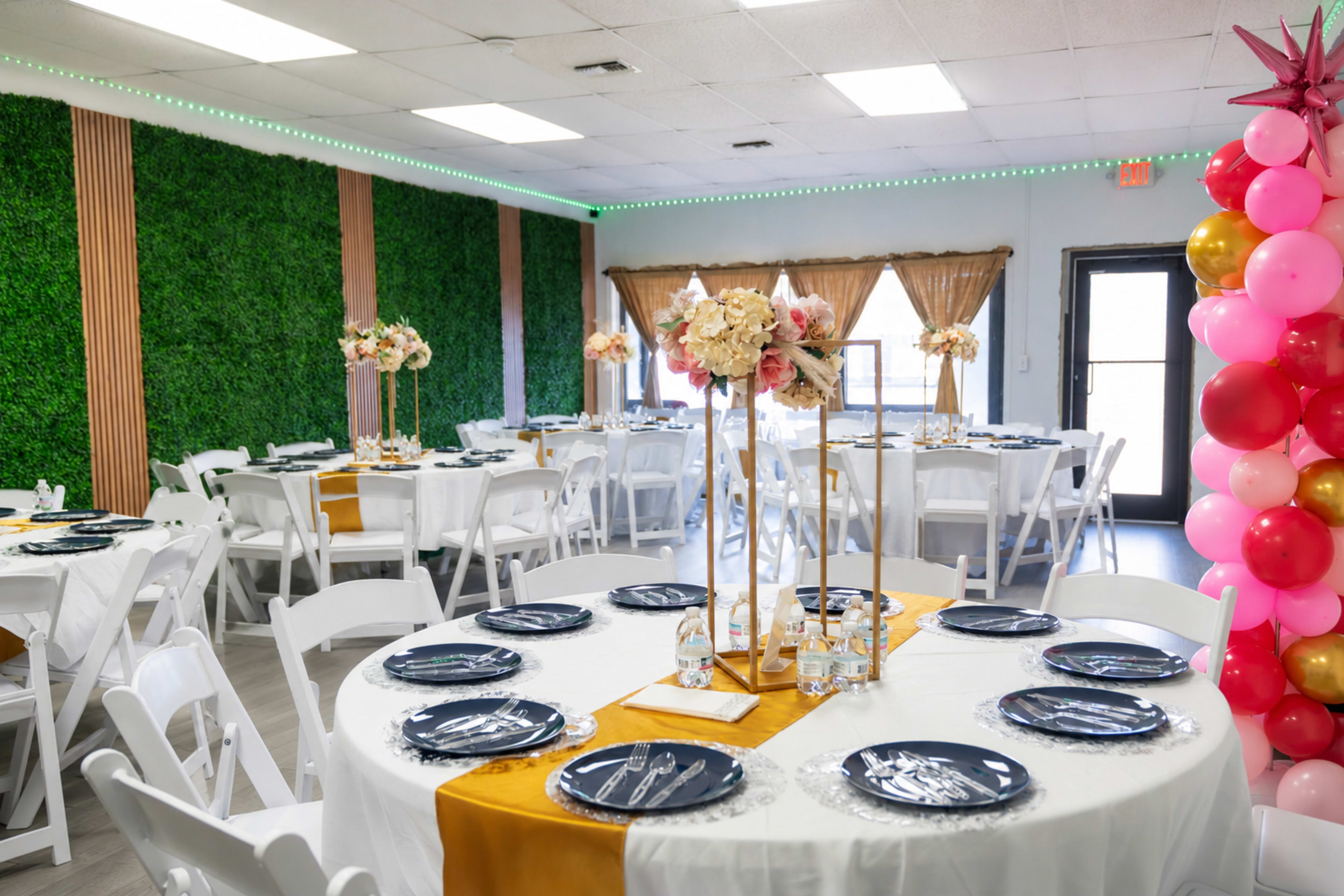 The image shows a banquet hall decorated with white tables set with navy blue plates and golden table runners, surrounded by greenery and colorful balloon arrangements.