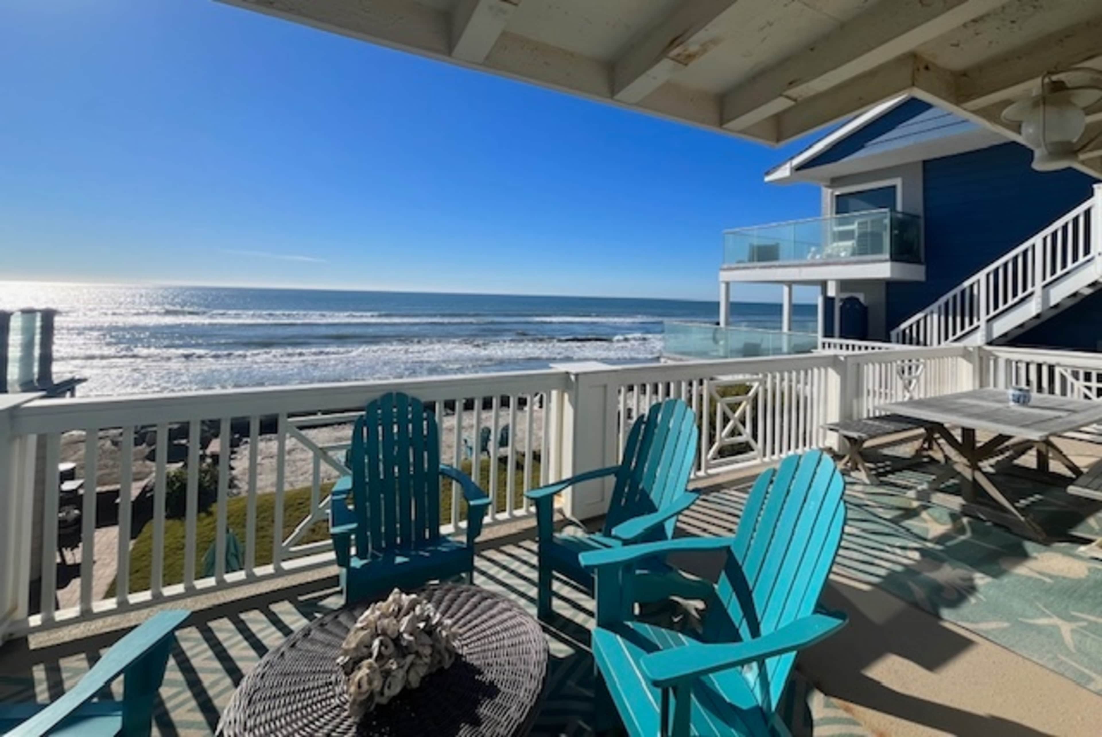 The image shows a beachfront patio with blue chairs overlooking the ocean and a house in the background.